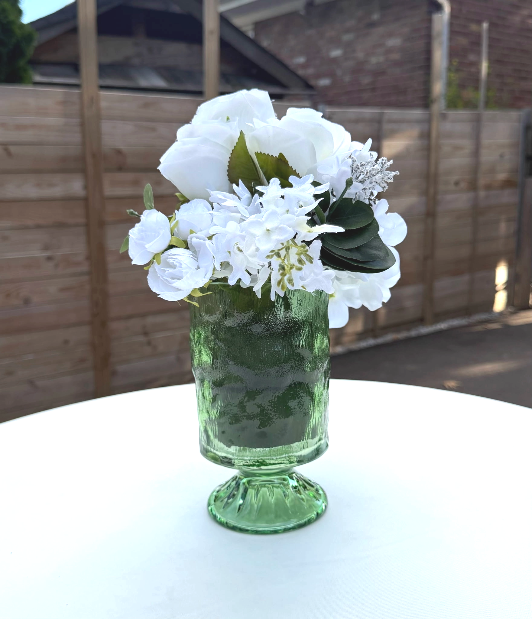 A green glass vase with a bouquet of white flowers sitting on a white surface, with a wooden fence and a brick building in the background.