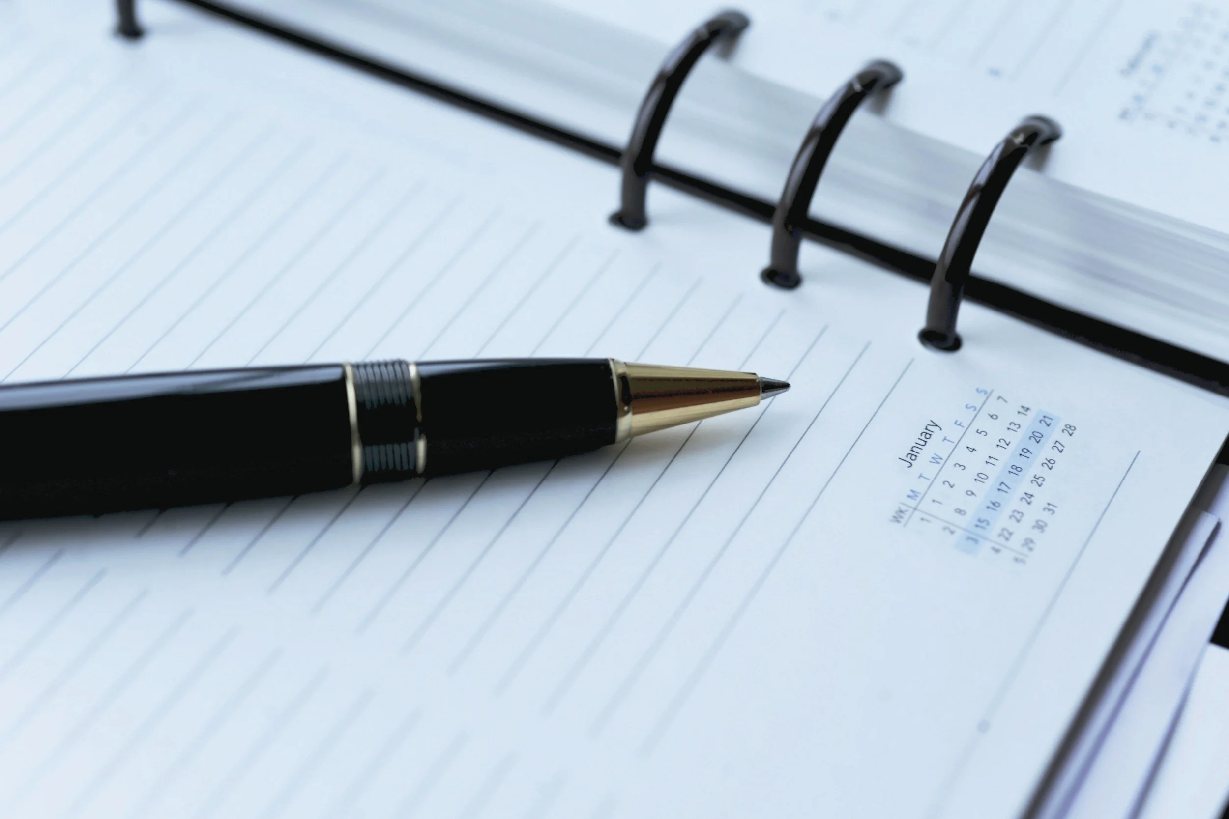Close-up of an open spiral-bound planner with a black and gold pen resting on the pages, and a calendar for January visible in the background.