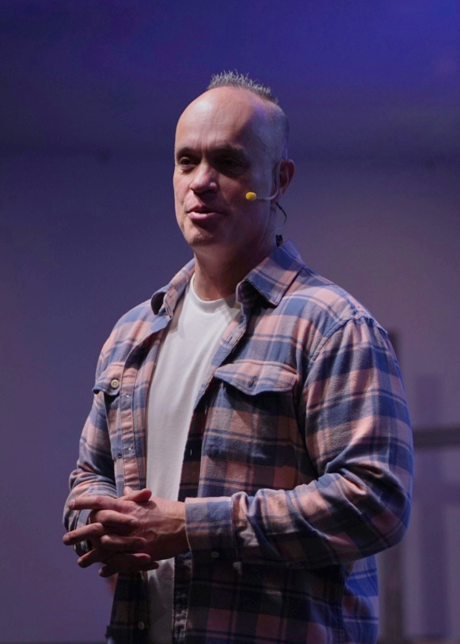 Man speaking on stage with a headset microphone, wearing a plaid shirt and white t-shirt, with a dark purple background.