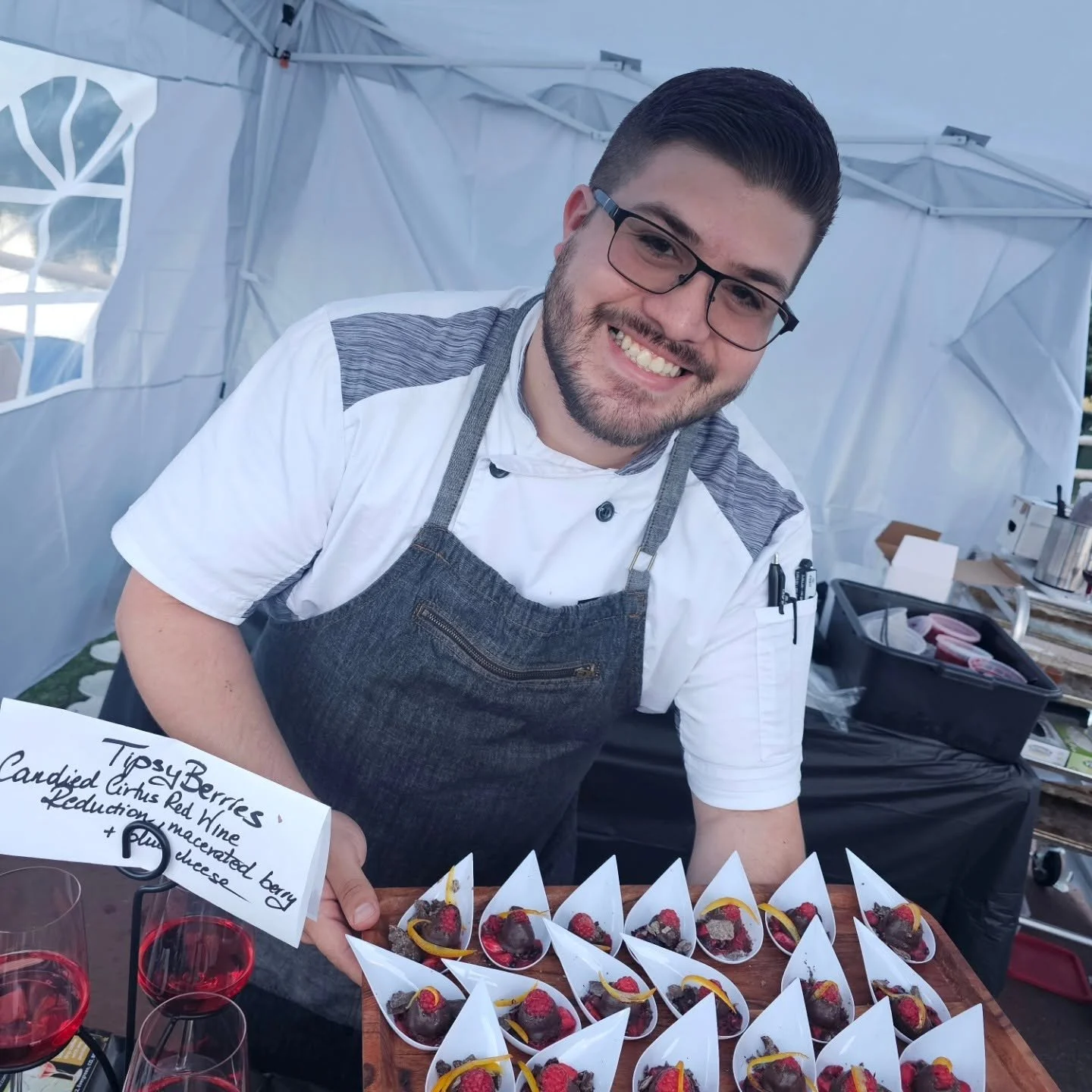 A smiling young man in a white shirt and gray apron at a food stall, presenting small chocolate dipped raspberries topped with candied orange