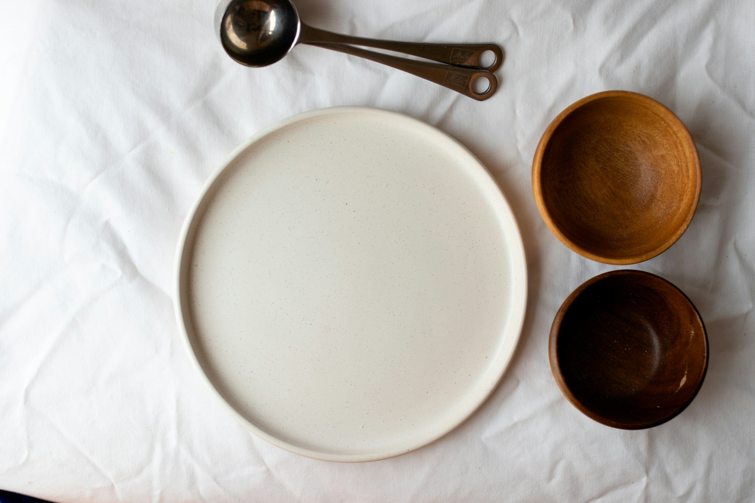 A white round ceramic plate on a white tablecloth, with a set of two brown measuring spoons above it, and two small brown ceramic bowls to the right.