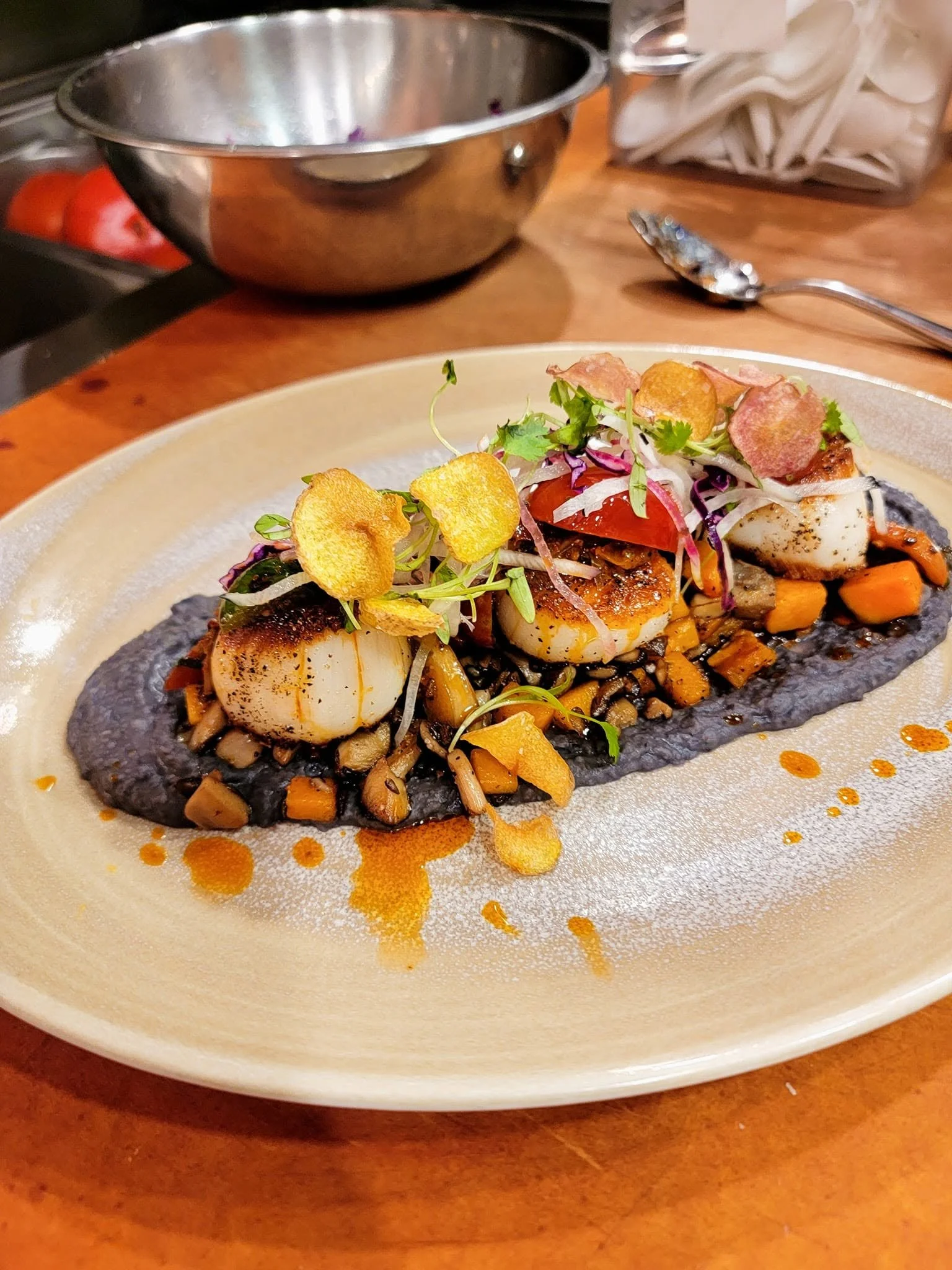 Seared scallops served on a bed of black bean puree with diced vegetables, garnished with microgreens and crispy potato chips, drizzled with a salsa matcha.