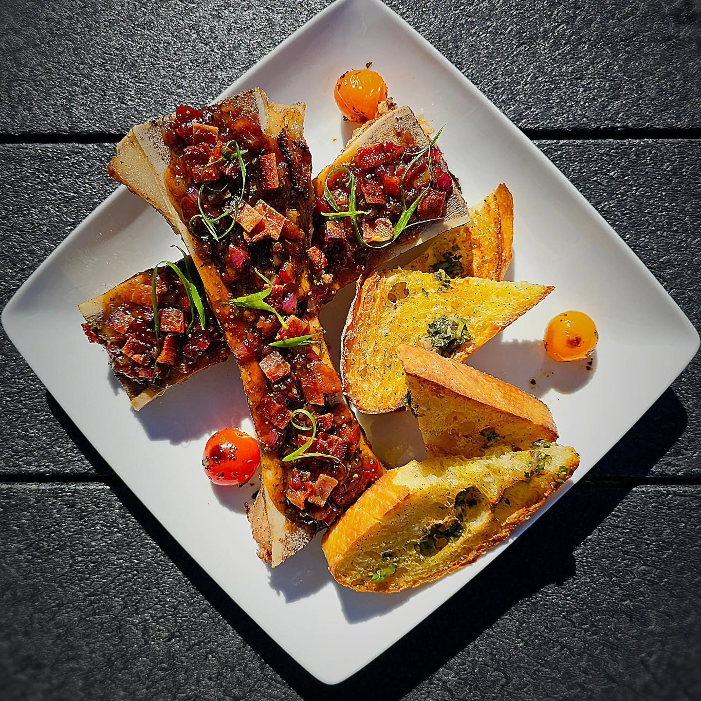 Seared Bone Marrow topped with a savory mixture of chopped onion jam and garnished with microgreens, and cherry tomatoes on a white square plate.