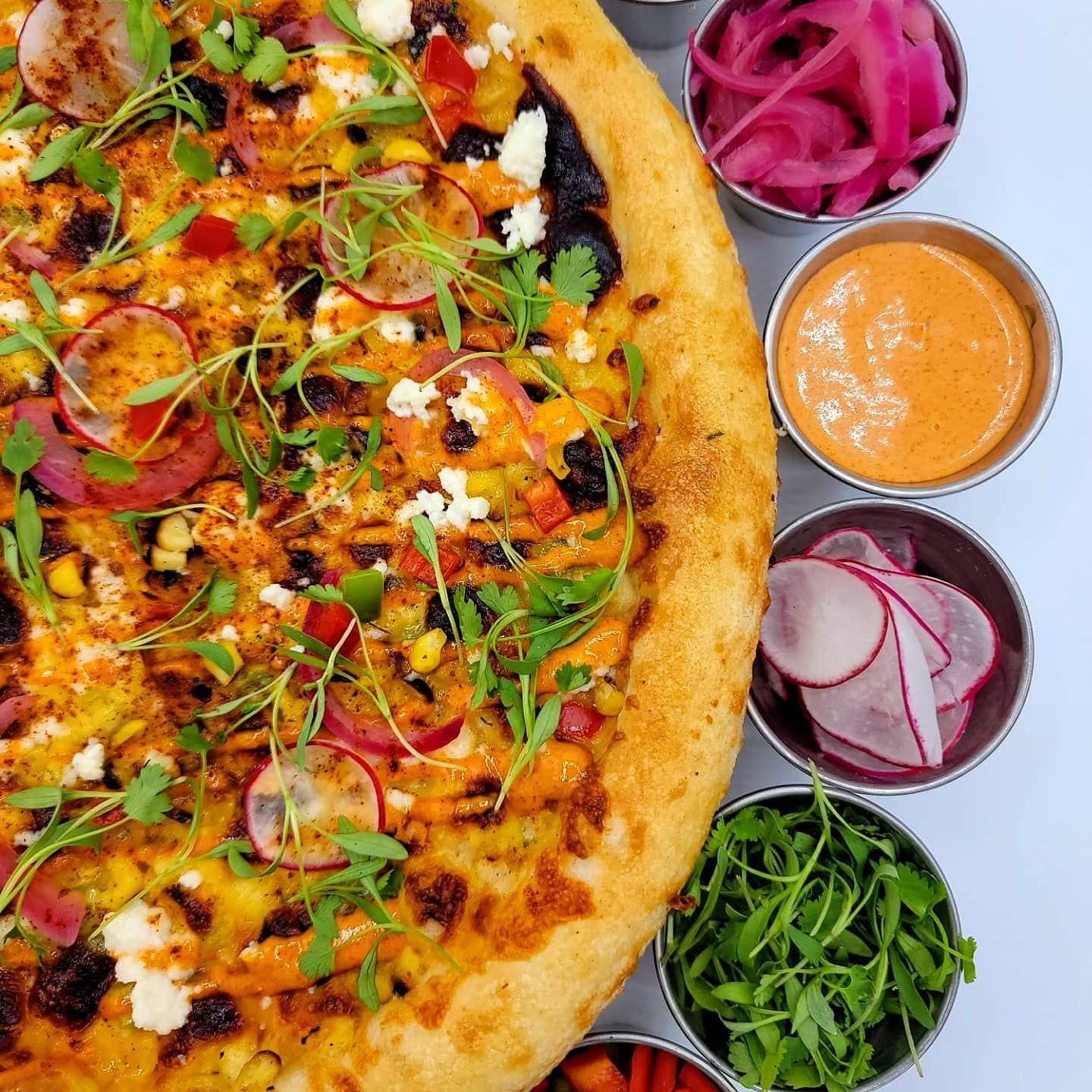 A pizza topped with cheese, sliced radishes, corn, black beans, chili flakes, and microgreens, served with side bowls of pickled onions, radish slices, sauce, and fresh cilantro.