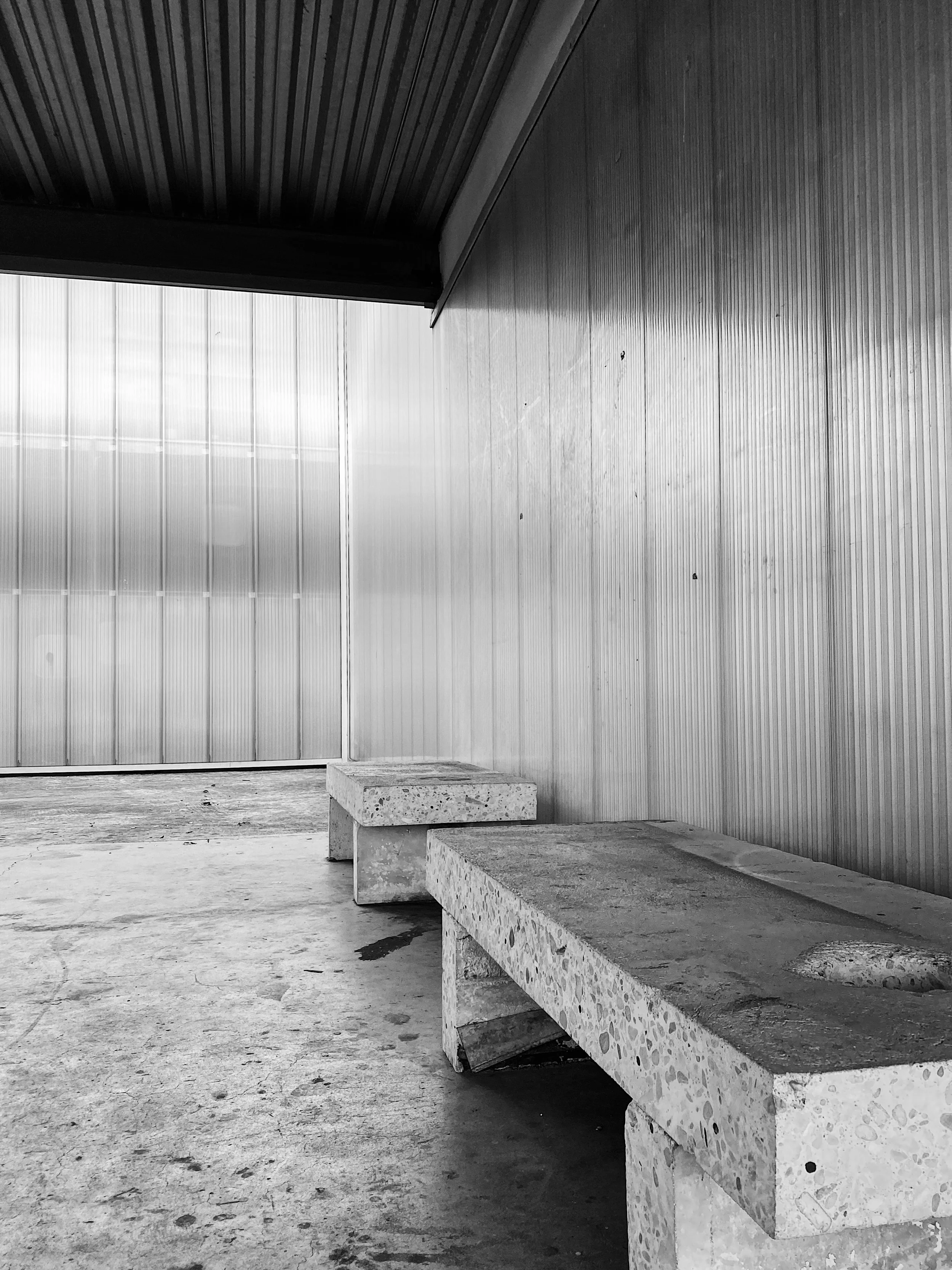 Black and white photo of a minimalistic modern space with two concrete benches, a vertical textured wall on the right, and a large window or translucent panel on the left, with a wooden or metal ceiling.