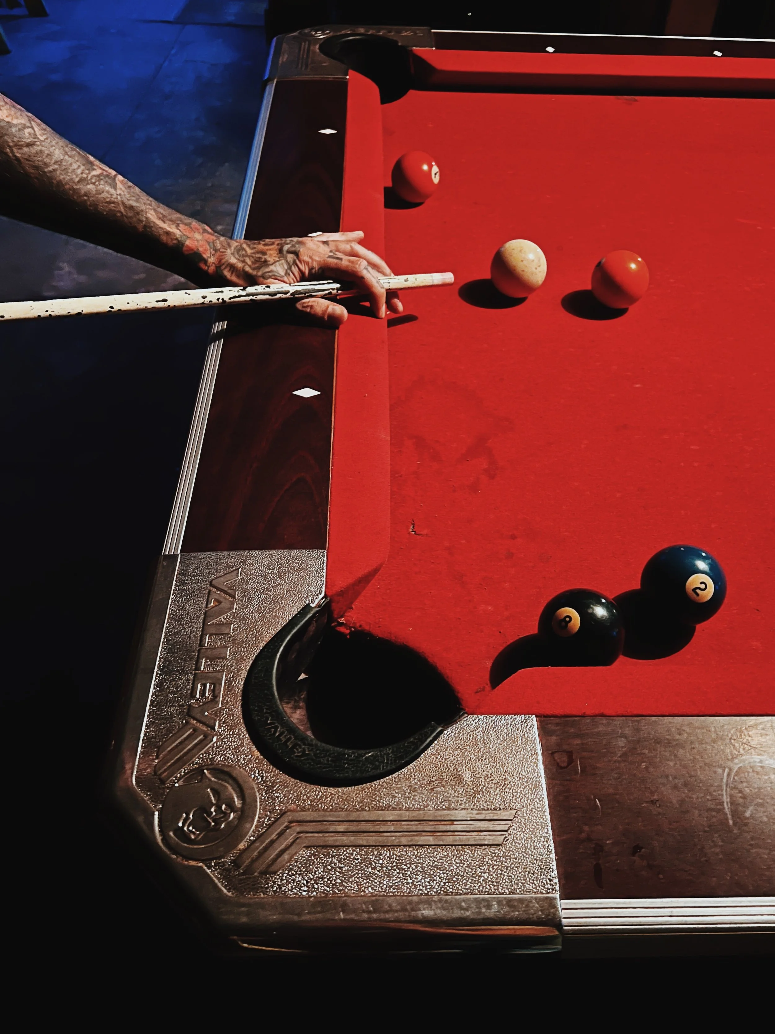 A person with tattooed arm taking a shot in a game of billiards, with a cue stick and billiard balls on a red table.