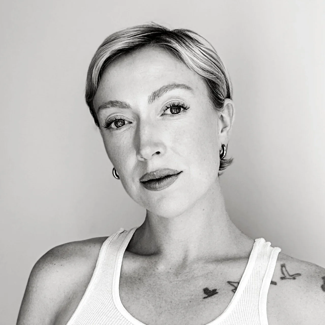 A black and white portrait of a woman with short hair, wearing earrings and a sleeveless top. She has a tattoo of birds on her shoulder and chest, and is looking directly at the camera with a slight smile.