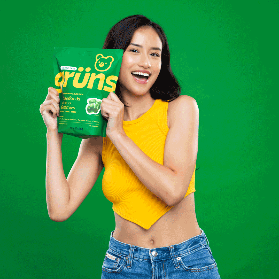 Young woman with long dark hair and a yellow crop top holding a green bag of gummies, smiling, against a green background.