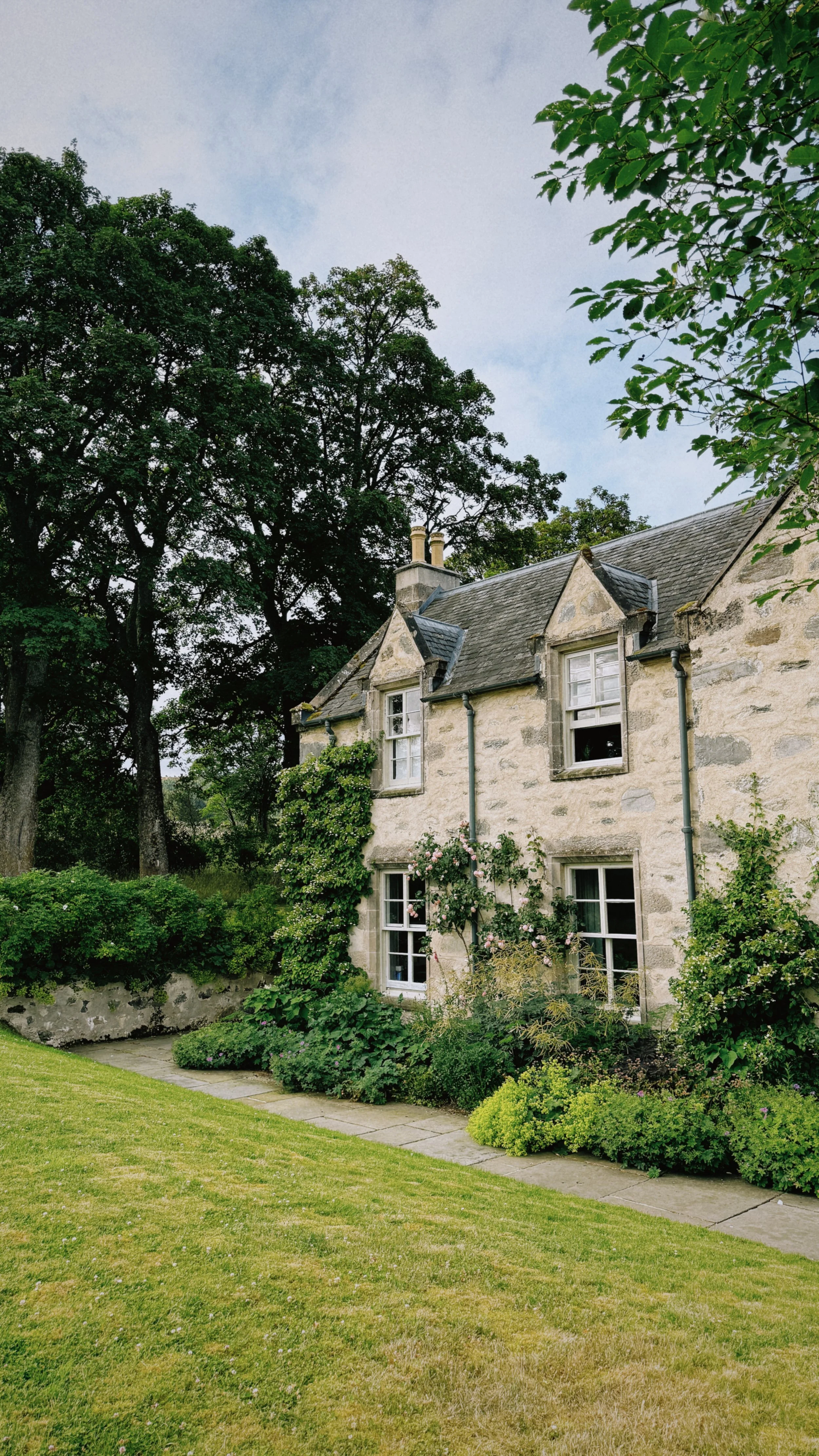 A stone cottage with a gray slate roof and multiple chimney stacks, surrounded by lush green trees and a well-maintained garden with shrubs and flowers.