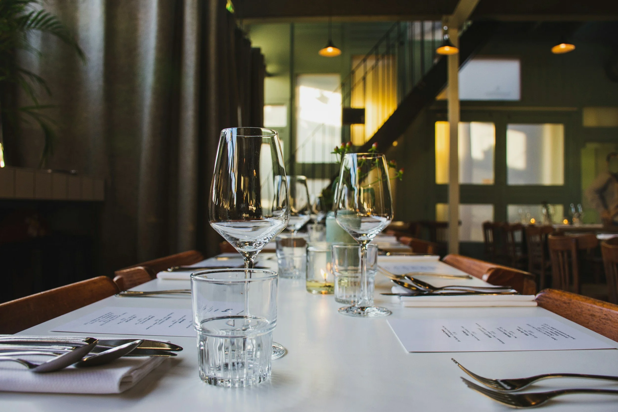 Set dining table with wine glasses, cutlery, and menus in a restaurant with large windows, green walls, and warm lighting.