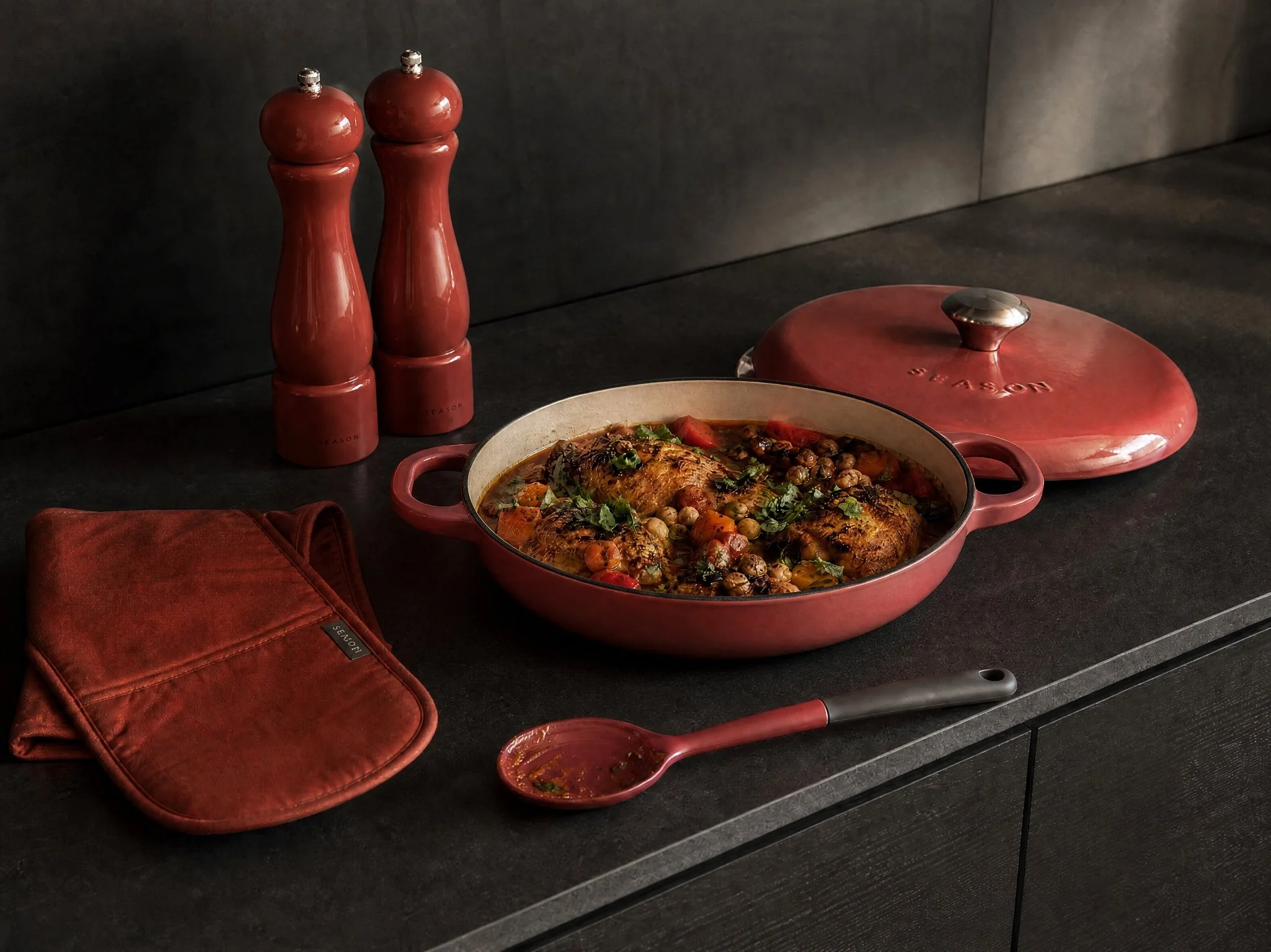 A red ceramic covered dish with food inside, topped with herbs and vegetables. Beside it, there is a spoon with sauce, a matching red oven mitt, and two tall red salt and pepper shakers on a black countertop against a dark background.