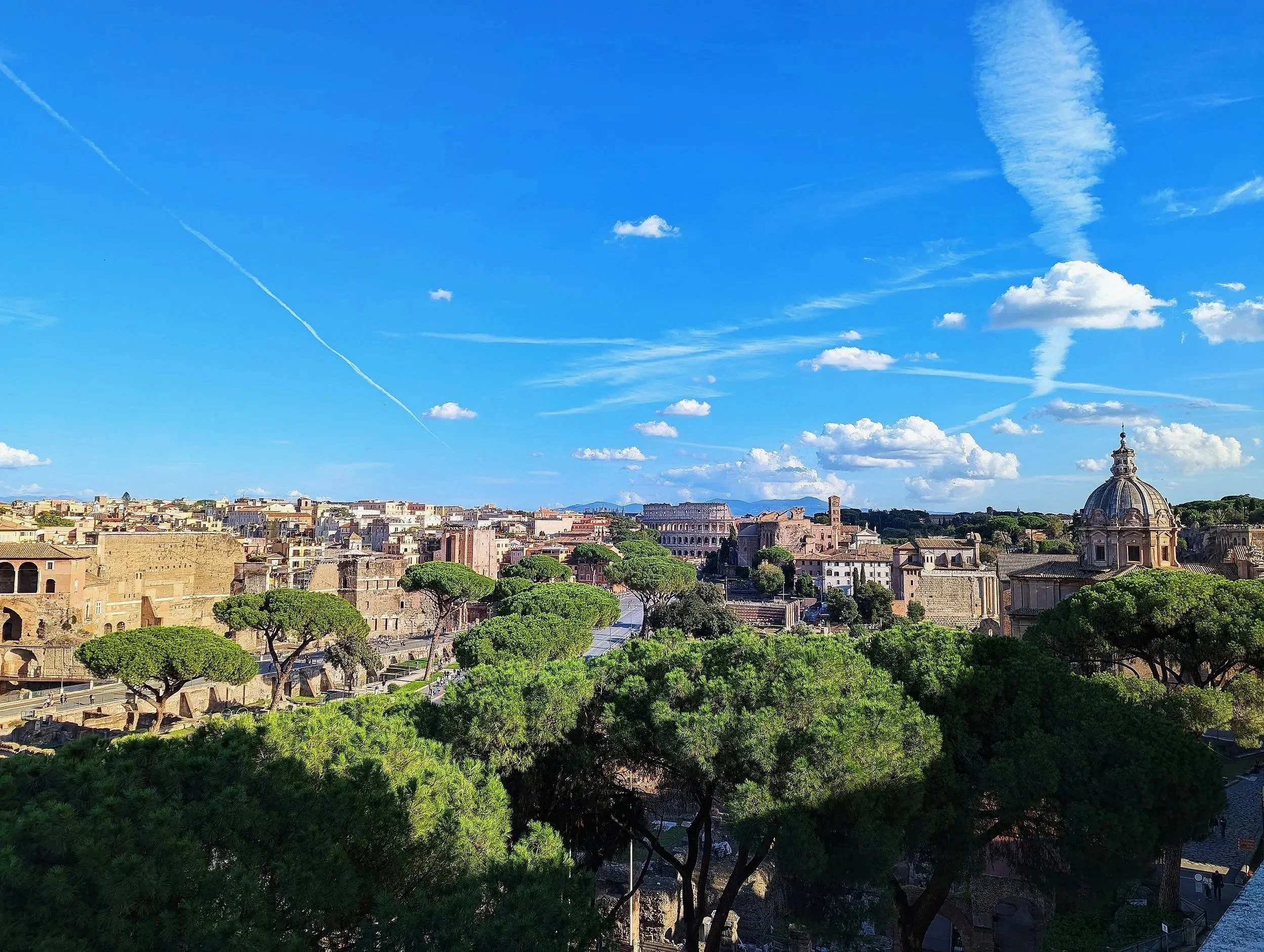 A panoramic view of Rome with ancient ruins, a large dome, and a cityscape under a bright blue sky with scattered clouds and contrails.