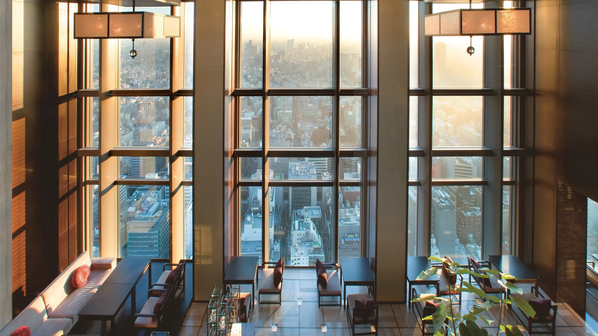 Interior view of a high-rise building with large floor-to-ceiling windows showing a cityscape skyline during sunset. The room features modern lighting fixtures hanging from the ceiling, a long wooden table, chairs, and a built-in seating area with cushions along the left side.