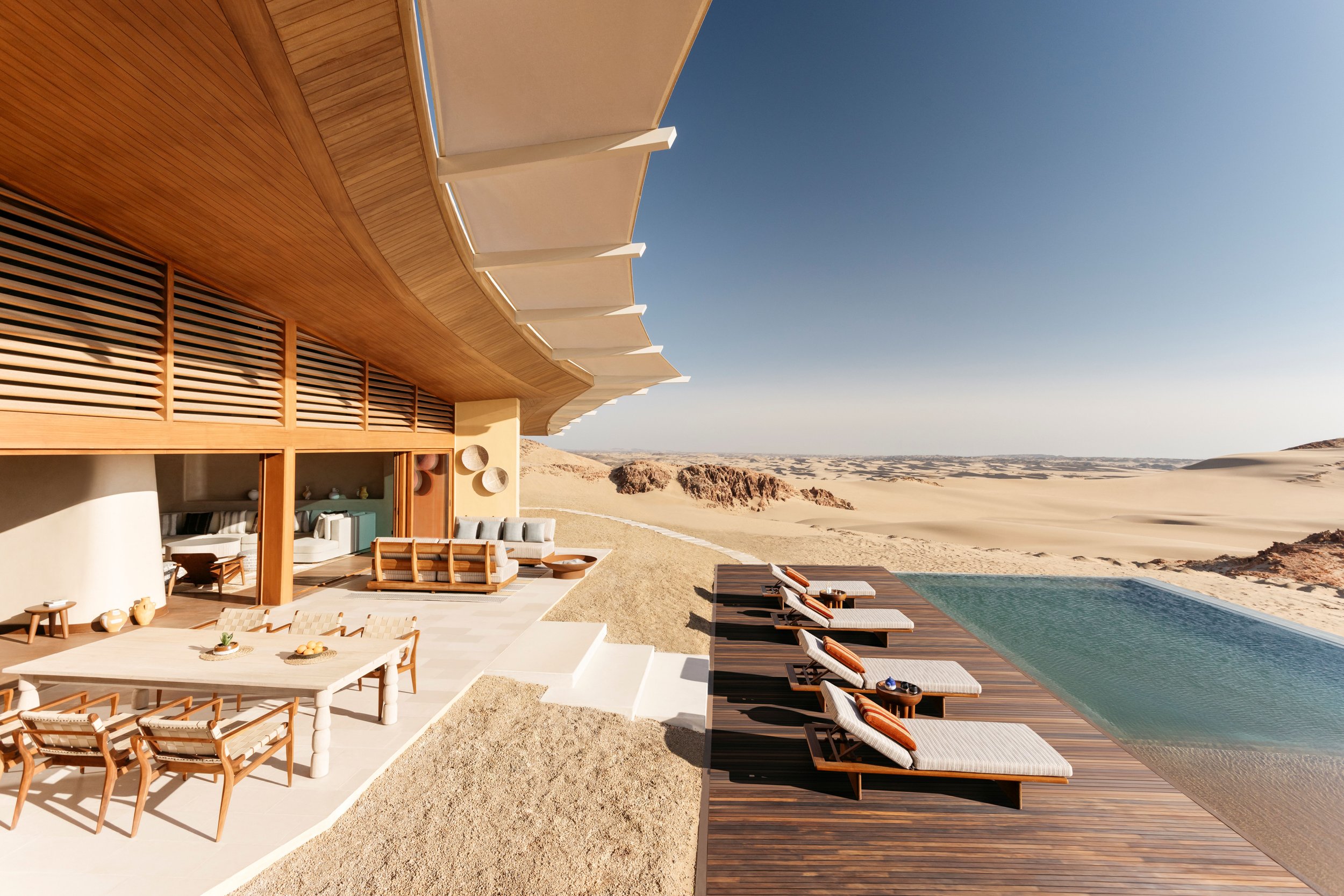 Modern house with wooden exterior and outdoor furniture overlooking a sandy desert landscape with dunes and rocks under a clear blue sky.