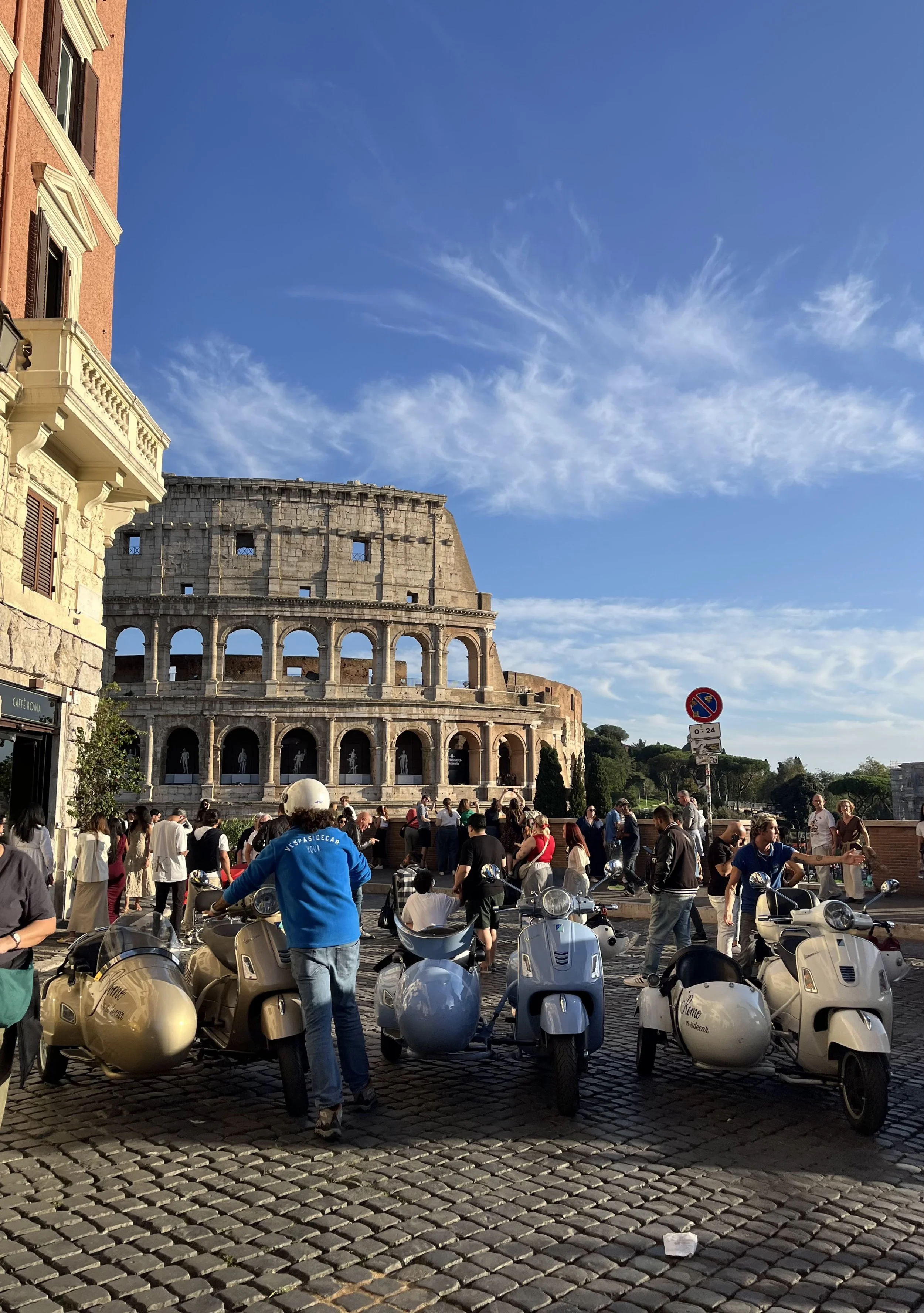 Tourists gathered around parked scooters in front of the Colosseum in Rome, Italy, under a blue sky with some clouds.