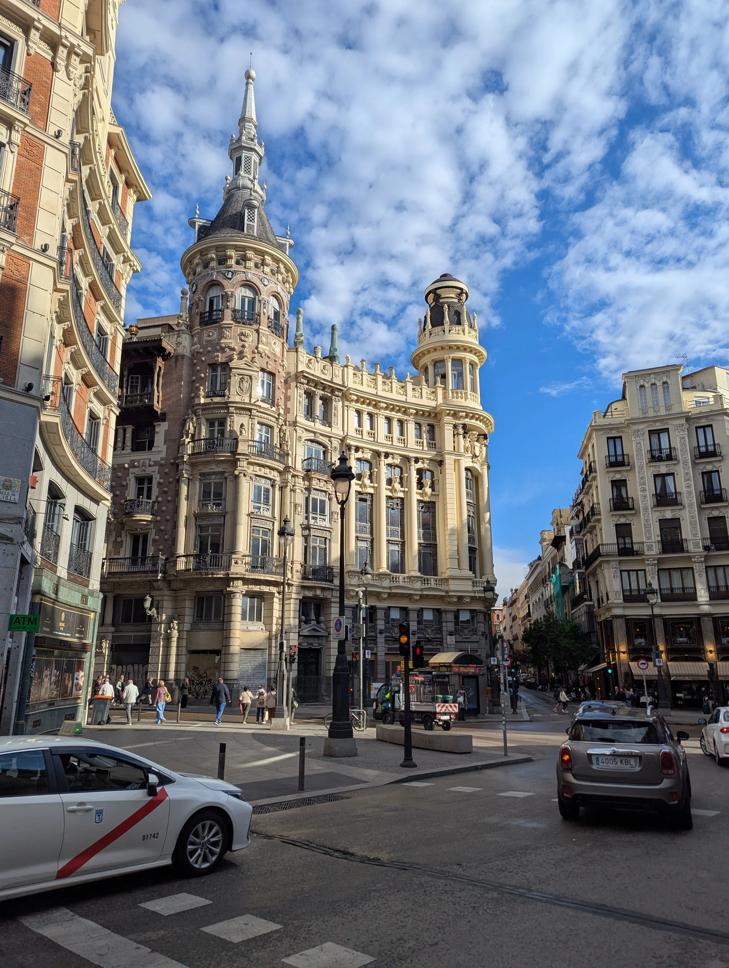 City street scene with historic buildings, people walking on sidewalk, cars on street, lampposts, and a partly cloudy sky.
