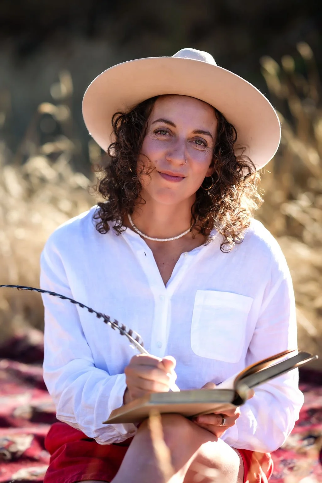 A woman with curly hair wearing a wide-brimmed hat, white shirt, and necklace, sitting outdoors, holding a notebook and pen, smiling at the camera.