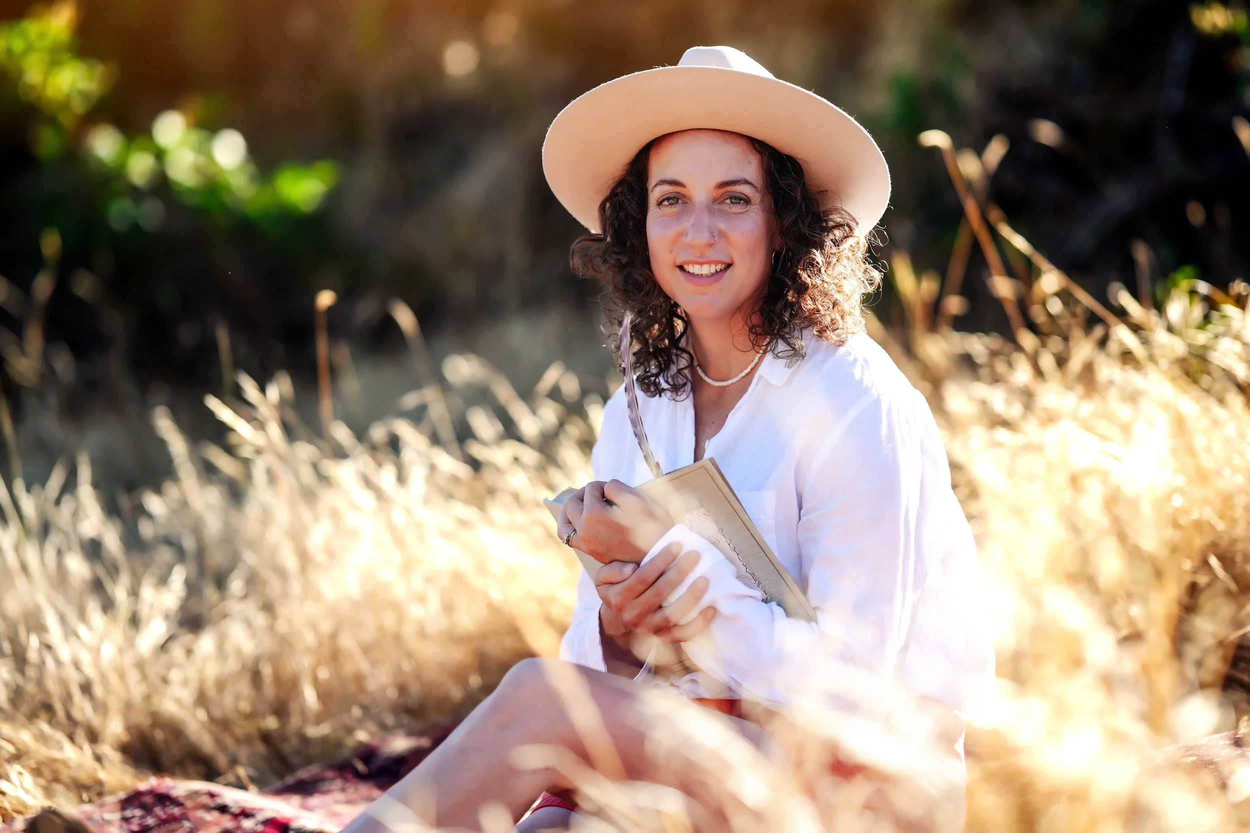 A woman with curly hair, wearing a white hat and white shirt, is sitting outdoors in a field of dry grass. She is smiling and holding a notebook and pen, with sunlight in the background.