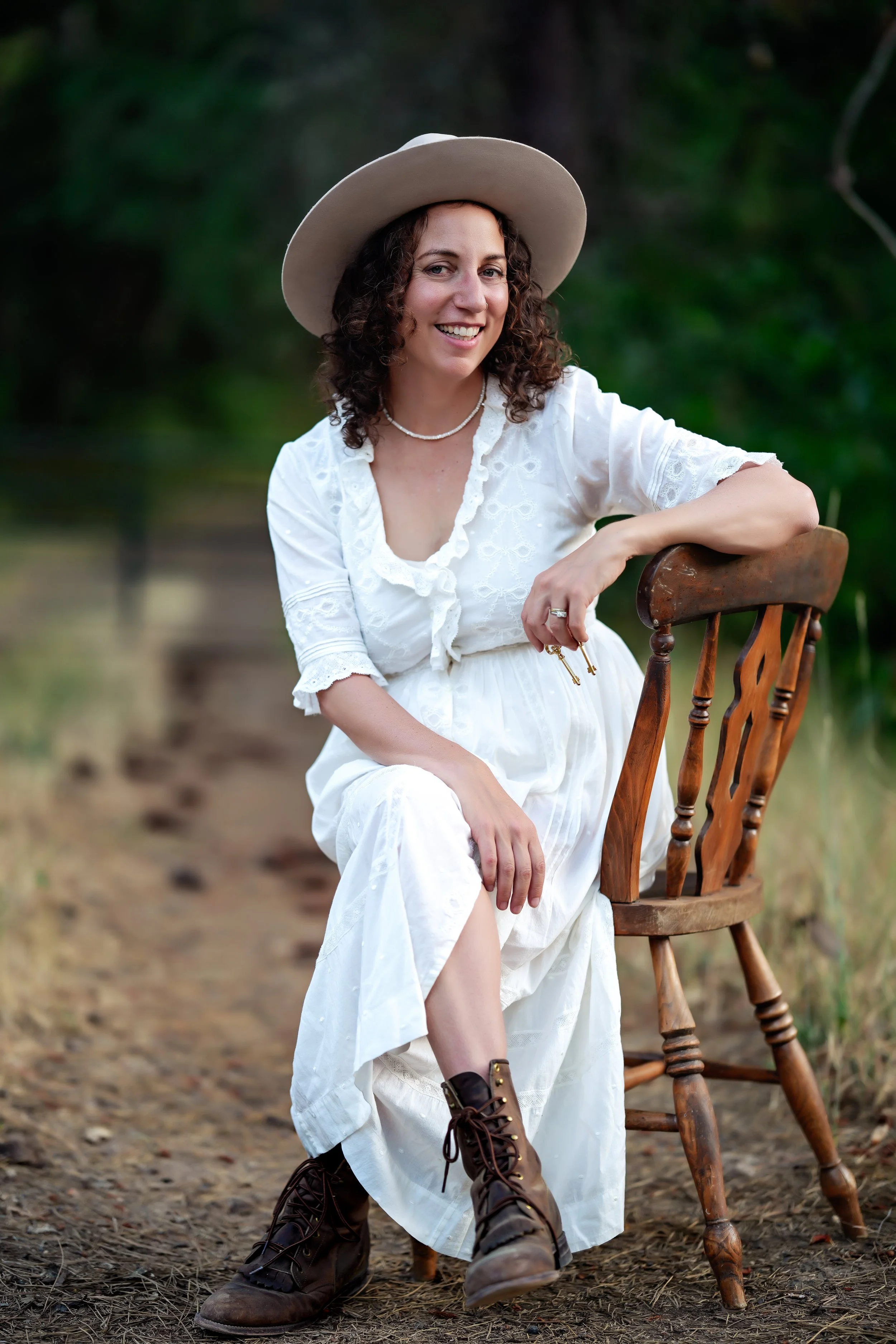 A woman with curly brown hair wearing a beige wide-brimmed hat, white dress with ruffled details, and brown lace-up boots, smiling while sitting outdoors on a wood chair, holding keys in her right hand, with a background of green trees and a dirt path.