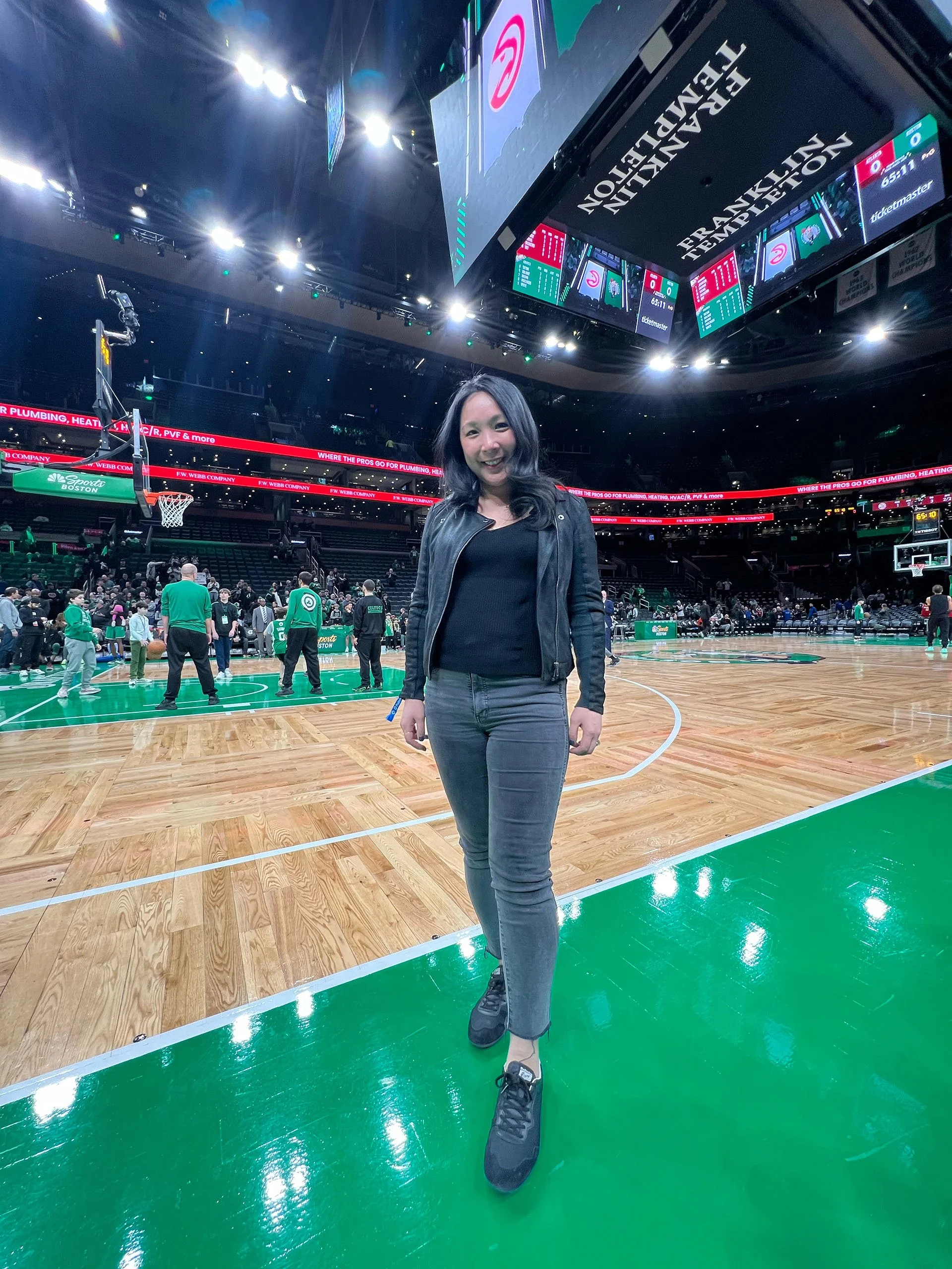A woman standing on a basketball court in an arena, smiling, with people and basketball hoops in the background. The arena has bright lights and large screens hanging from the ceiling.