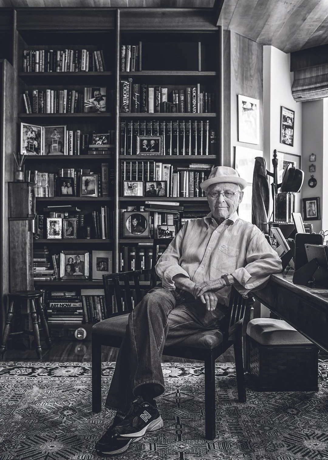 An elderly man wearing a hat, glasses, and casual clothes sitting in a wooden chair in a cozy home library, surrounded by books and framed photographs with a window behind him.
