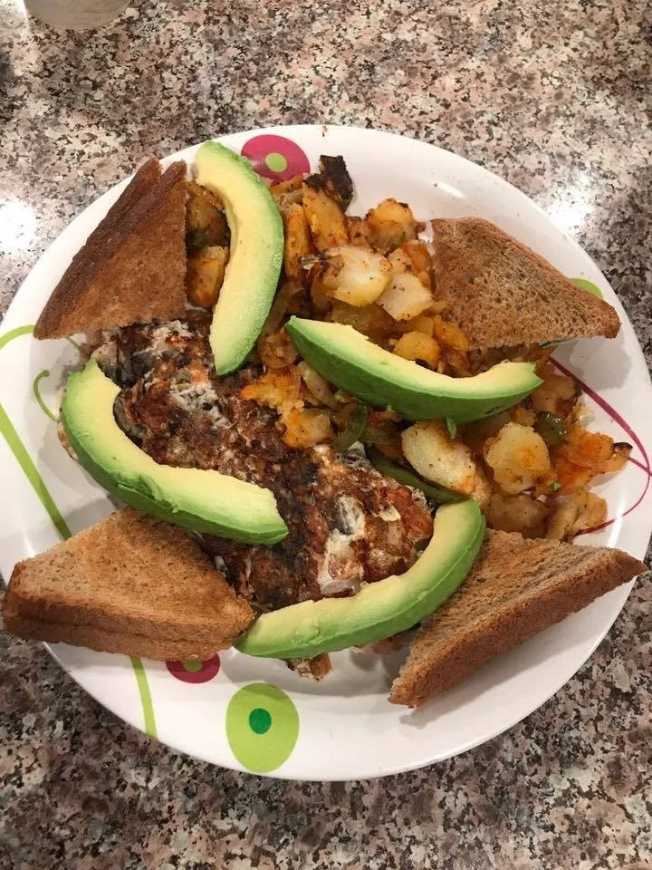 Plate of breakfast featuring fried eggs topped with melted cheese, sliced avocado, toasted bread, and diced potatoes.