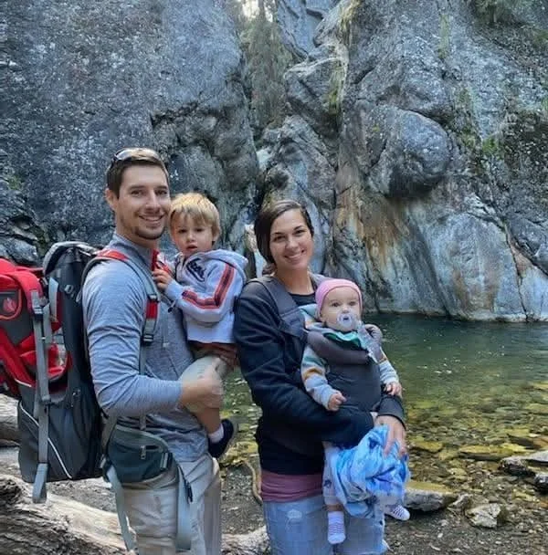 Family of four, a father, mother, and two young children, standing by a creek in a rocky outdoor setting during daytime. The father carries a child with a pacifier, while the mother holds a baby.
