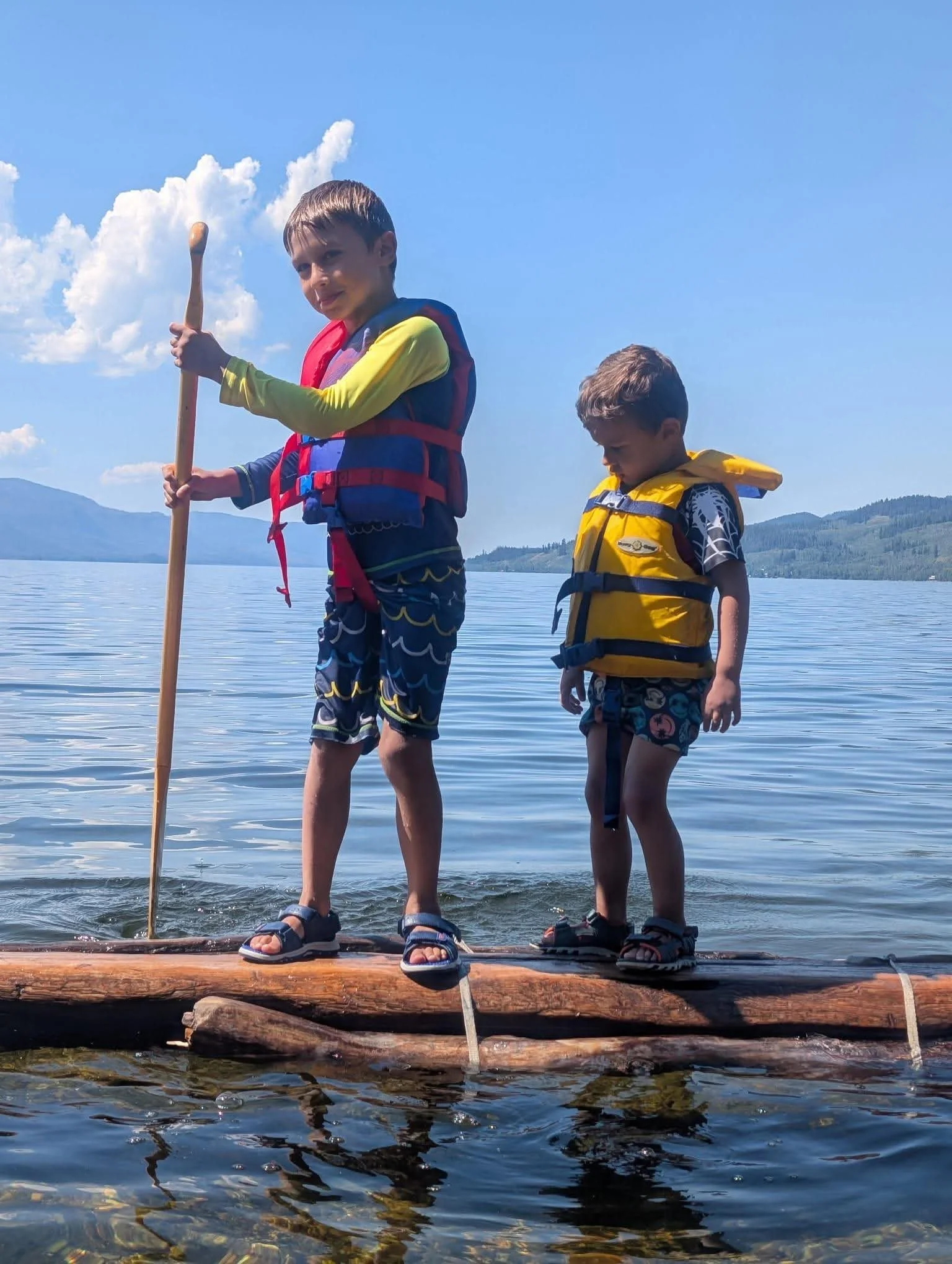 Two young boys standing on a log float at the edge of a lake, wearing life jackets and sandals, with a mountain and blue sky in the background.