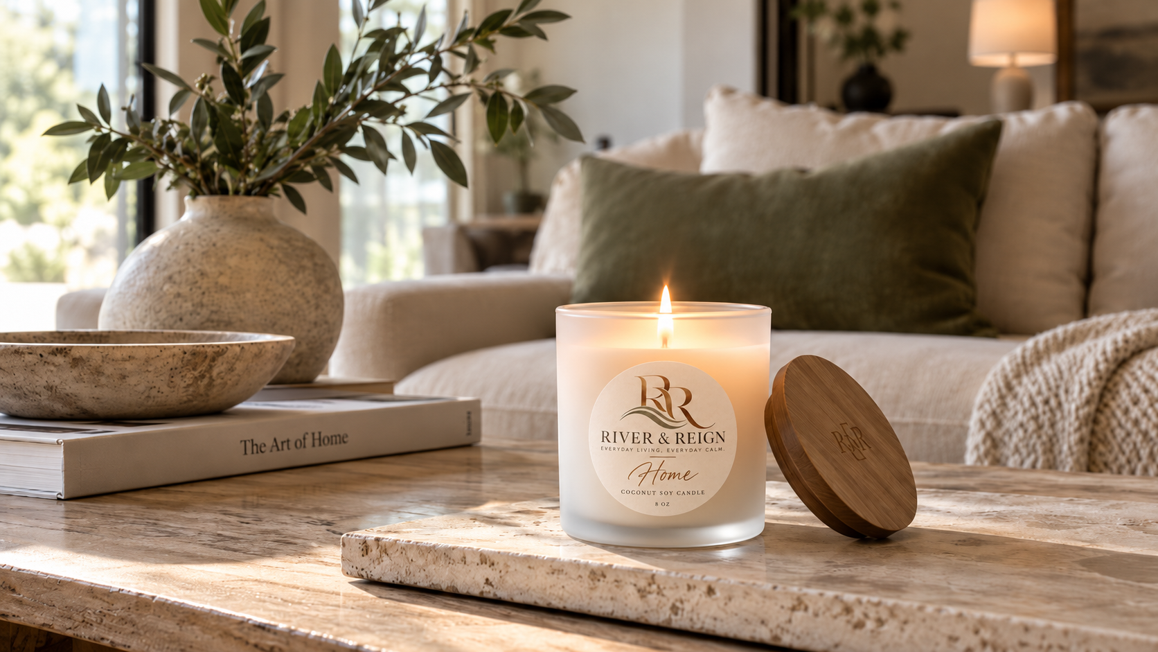 A lit River & Reign candle on a marble coffee table beside a cream sofa in a cozy, modern living room.
