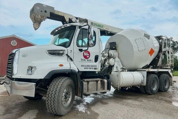 A white concrete mixer truck with a large drum and a boom lift parked on a muddy surface near a red barn. The truck has markings indicating it belongs to a concrete company.