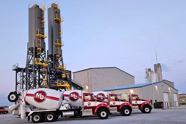 Three red cement mixers parked outside an industrial building with tall silos and a cloudy sky.