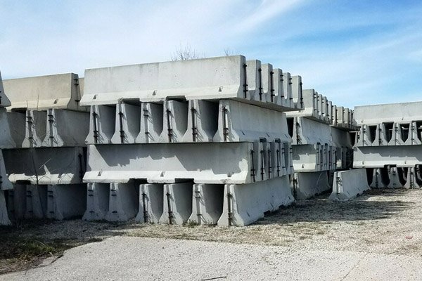 A stack of large concrete barriers outdoors on a gravel surface under a partly cloudy sky.
