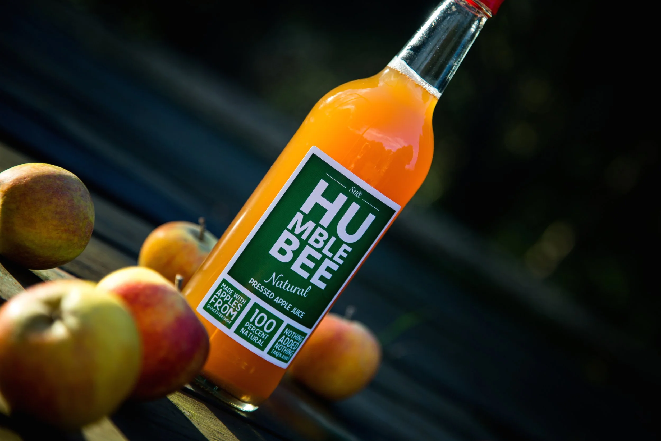 A glass bottle of natural pressed apple juice labeled 'Humble' surrounded by fresh apples on a wooden surface outdoors.