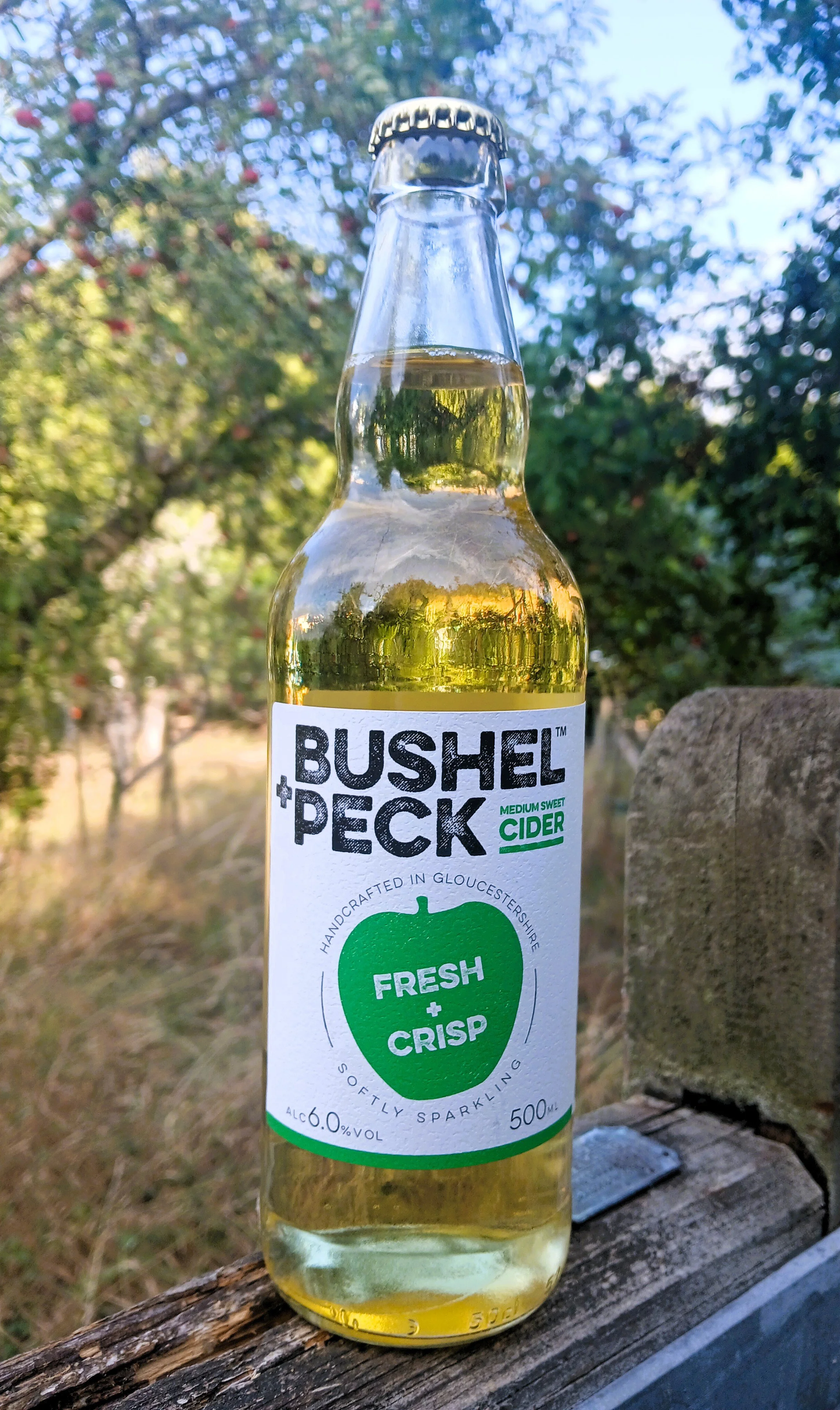 A bottle of Bushel Peck medium sweet cider placed on a wooden surface outdoors, with trees and blue sky in the background.