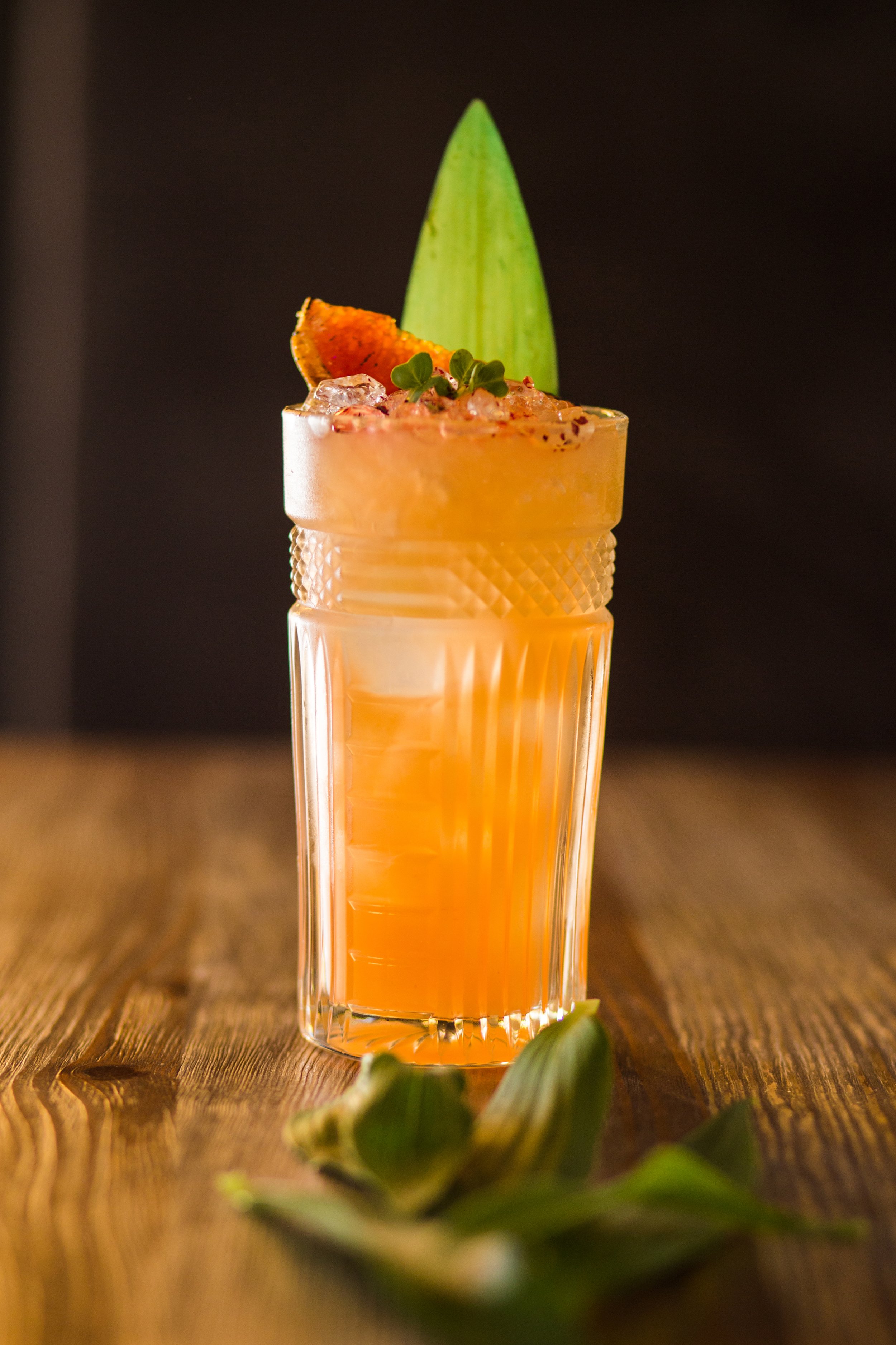 A tall glass of orange cocktail garnished with a slice of orange, microgreens, and a pineapple leaf in a dark background, placed on a wooden surface.