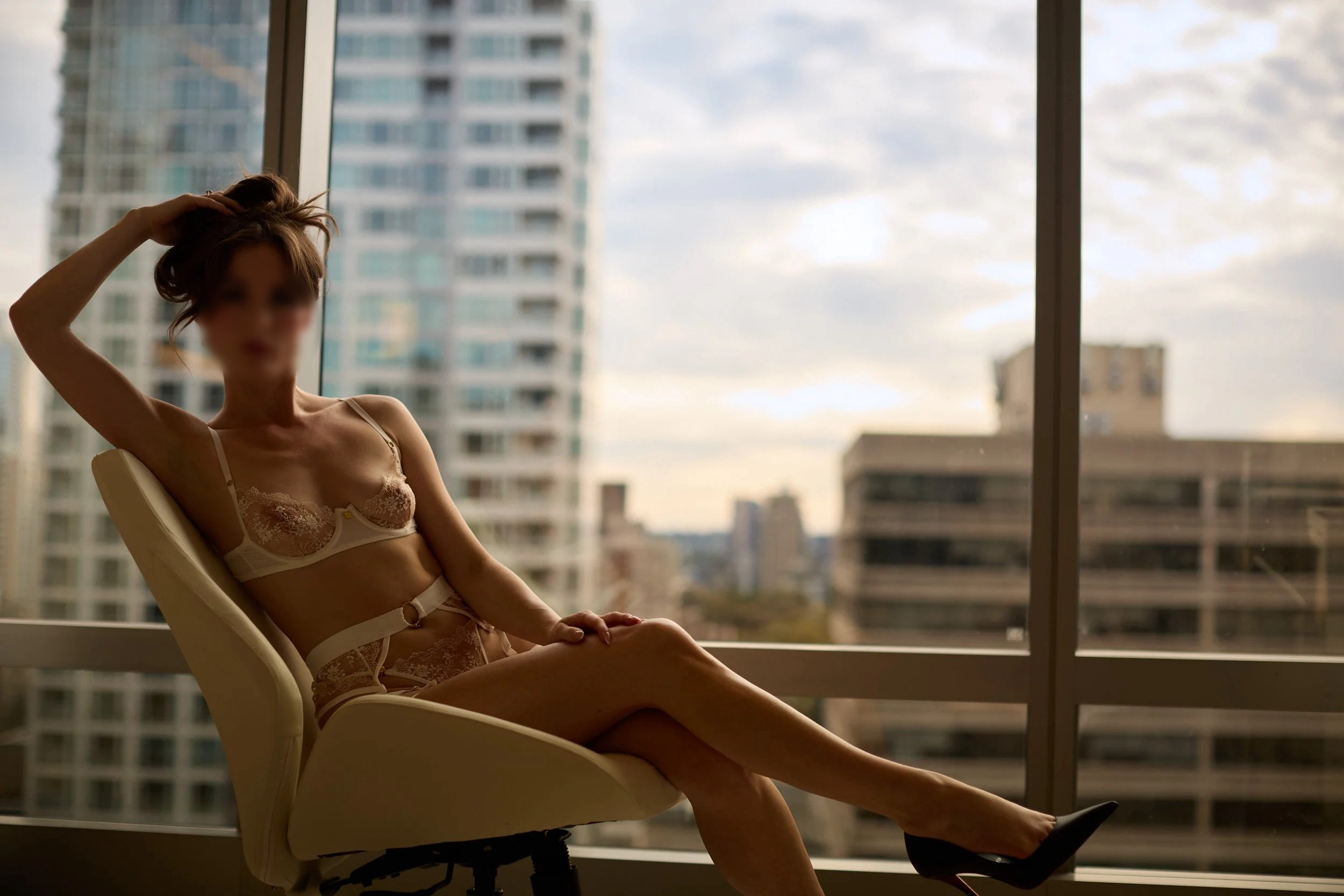 A woman in lace lingerie sitting on a white chair in front of a large window with a cityscape view of tall buildings and a cloudy sky.