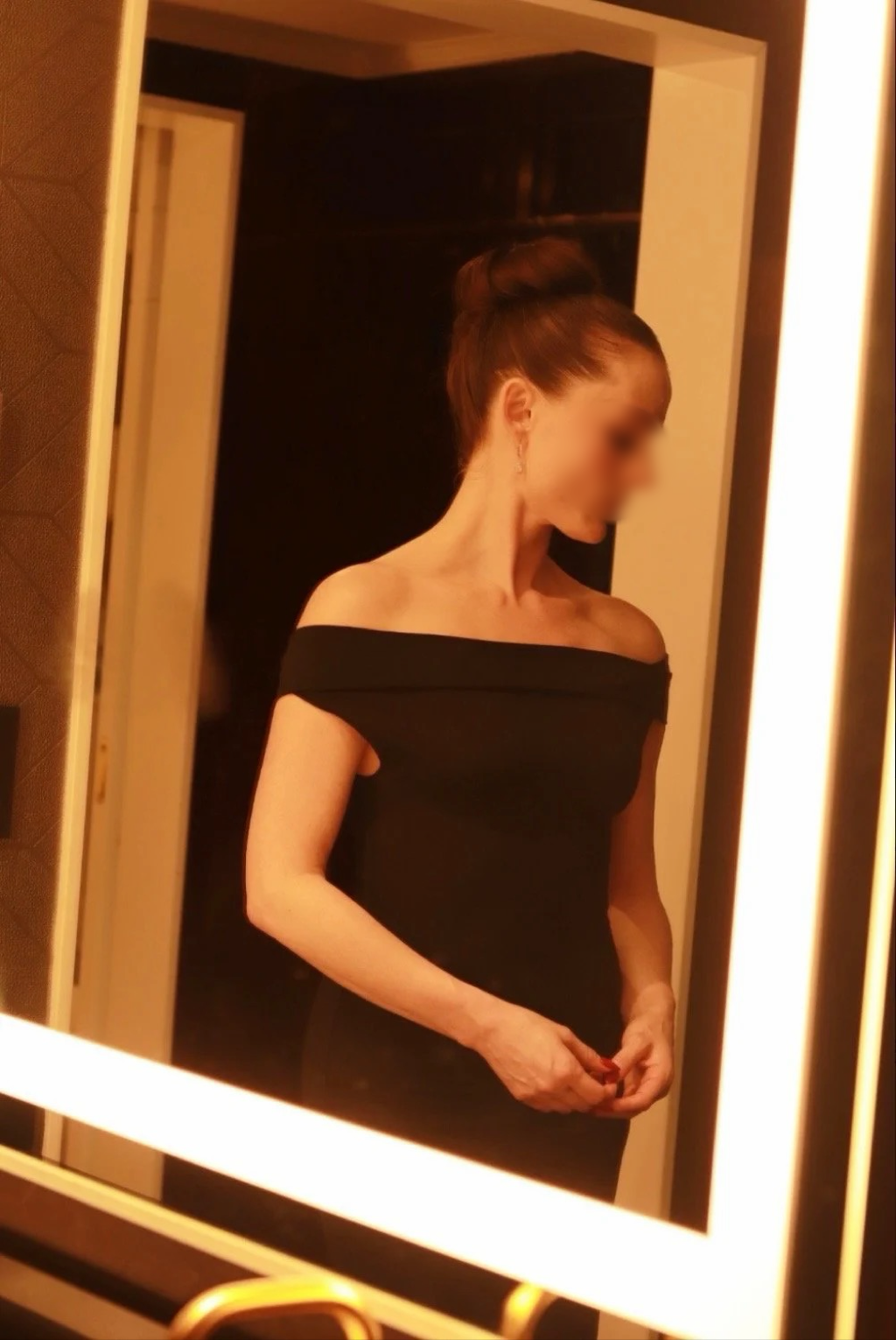 A woman in a black off-the-shoulder dress looking at herself in a mirror with warm lighting.