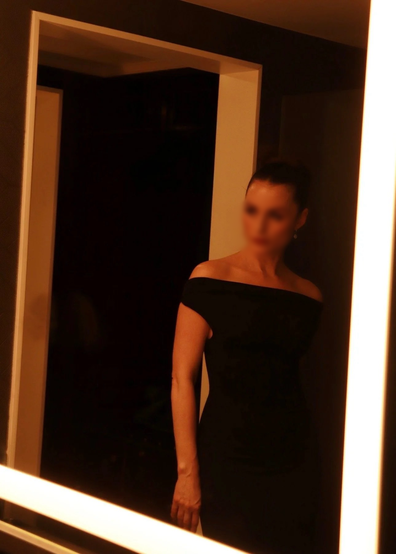 A woman in a black off-shoulder dress standing in front of a mirror, with her face blurred.