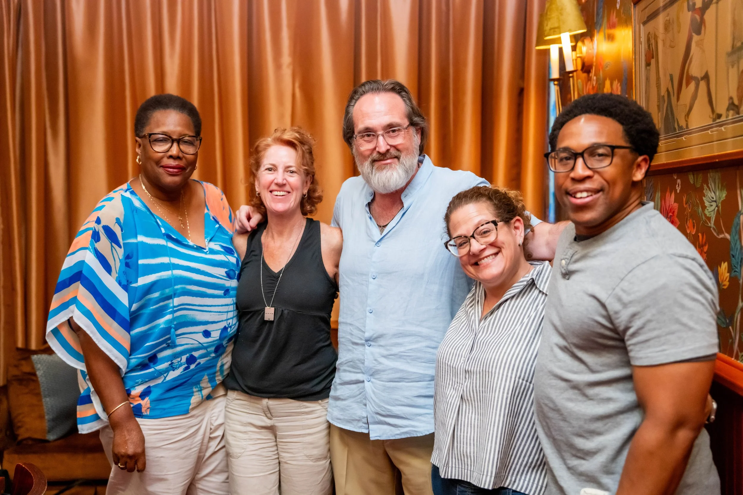 Group of six diverse adults smiling and posing together indoors, with a warm orange curtain and floral artwork in the background.