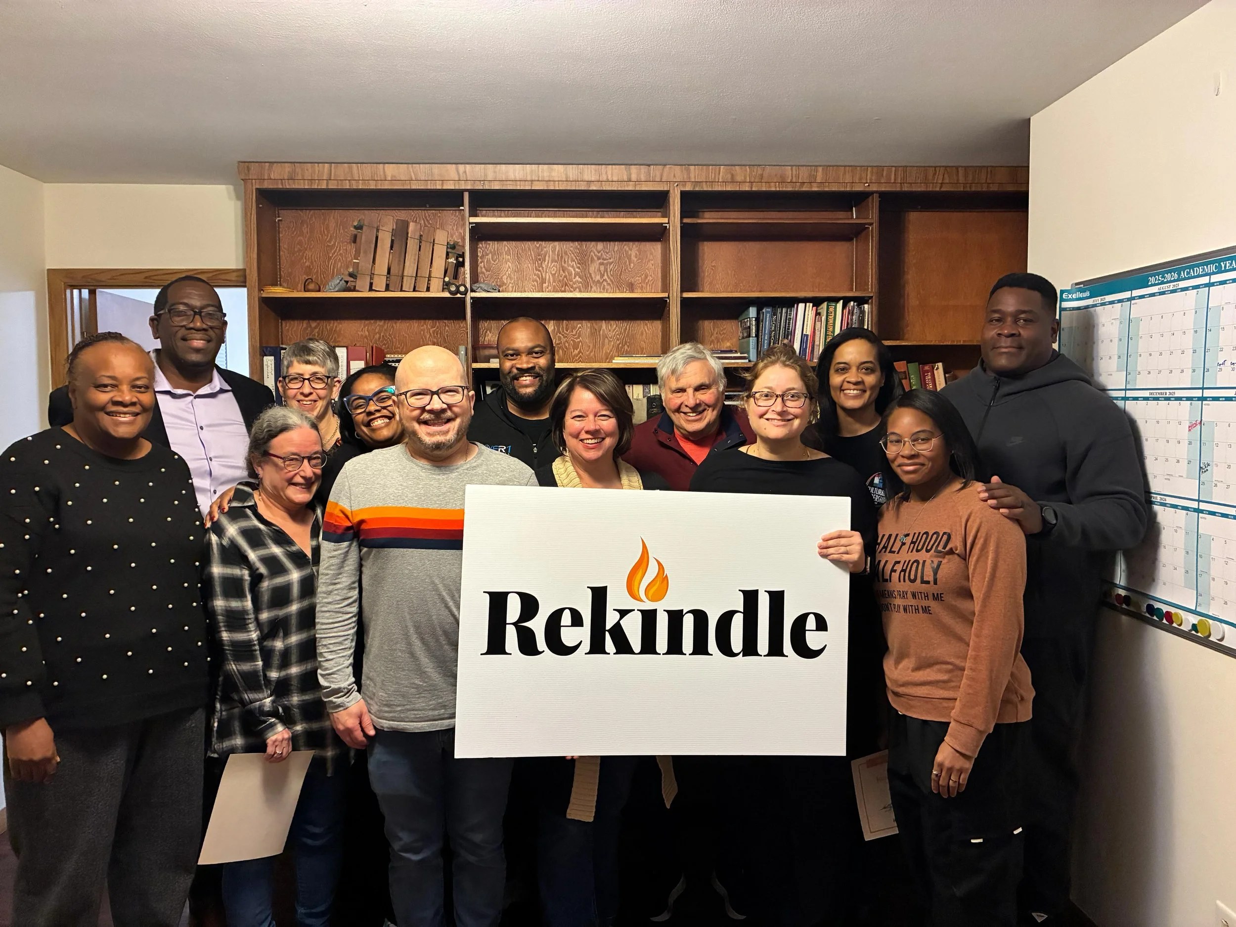 Group of diverse smiling people standing indoors, holding a sign that reads "Rekindle" with a flame symbol, in front of wooden bookshelves.