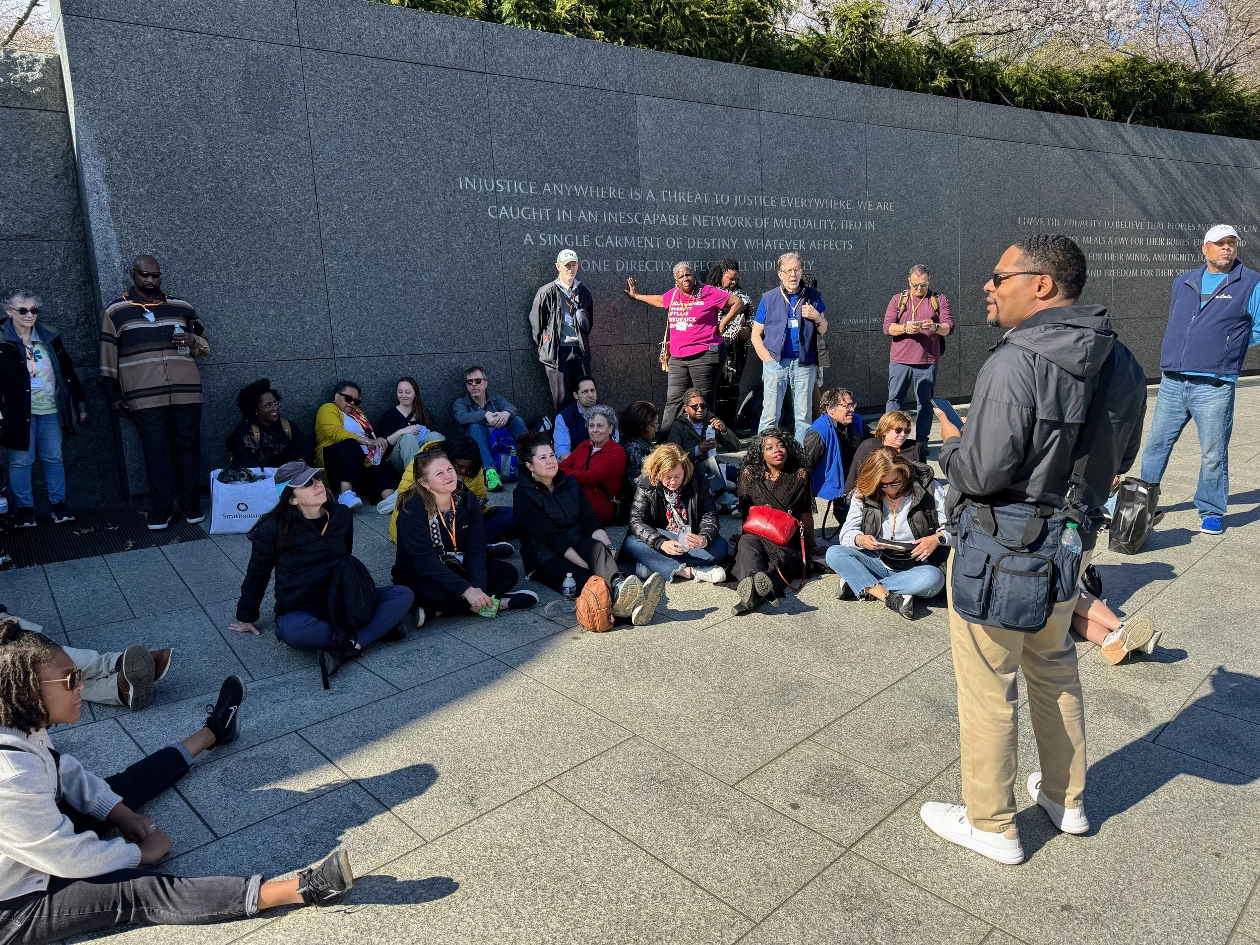 A group of people sitting and standing outdoors against a dark gray wall, with one person speaking to the group. The wall has an inscription about justice.