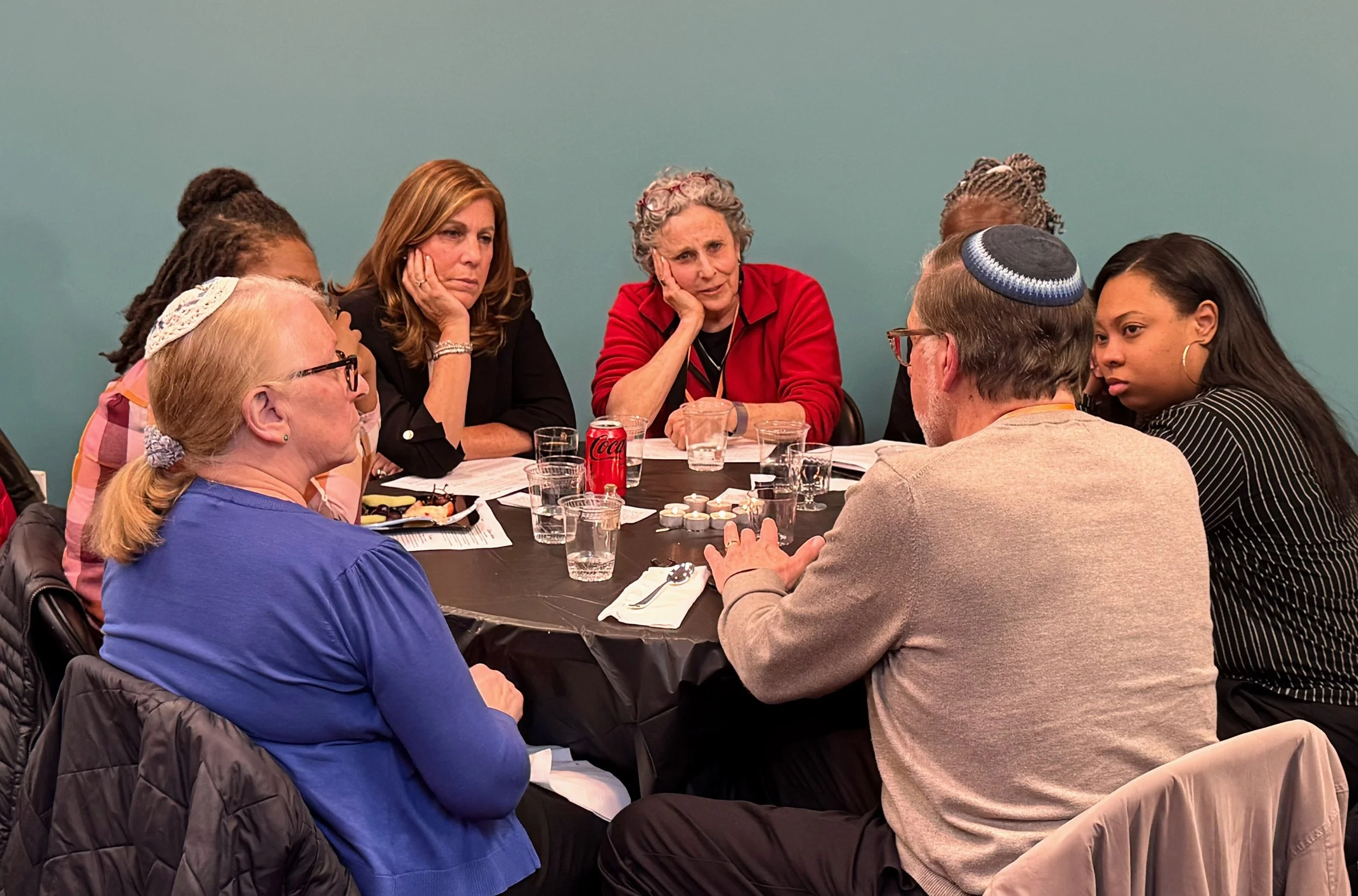 A group of diverse people sitting around a covered dinner table, engaged in conversation in a casual setting.