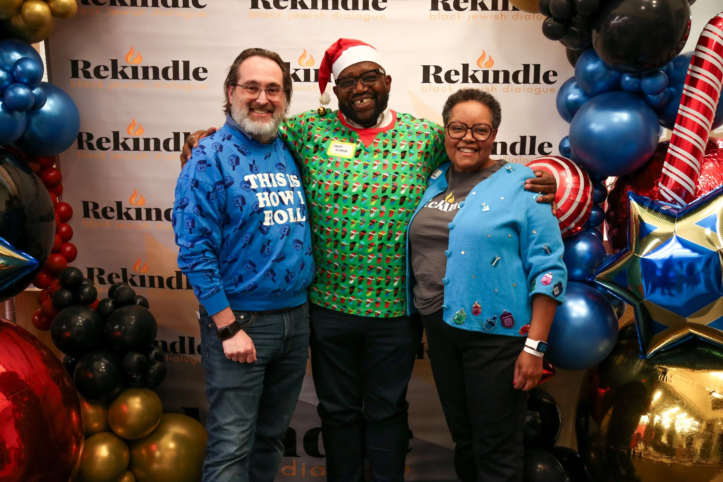 Three people smiling and standing together in front of a white backdrop with 'Rekindle' text and a flame logo, surrounded by colorful balloons, during a holiday celebration. The person in the middle is wearing a Santa hat and a festive sweater, while the person on the left wears a disposable blue gown with white text, and the person on the right wears glasses and a light blue jacket with pin decorations.