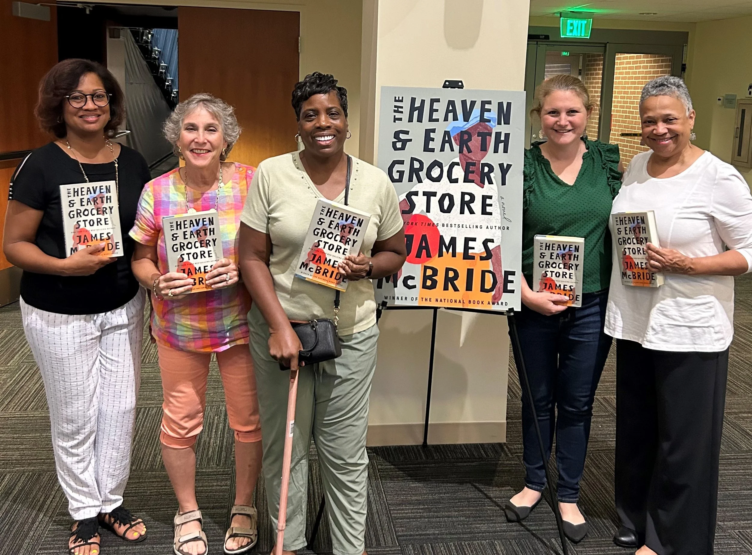 A group of five women standing indoors, holding copies of a book titled 'The Heaven & Earth Grocery Store' by James McBride. There is a sign in the background with the same book title and a rainbow-colored cover.