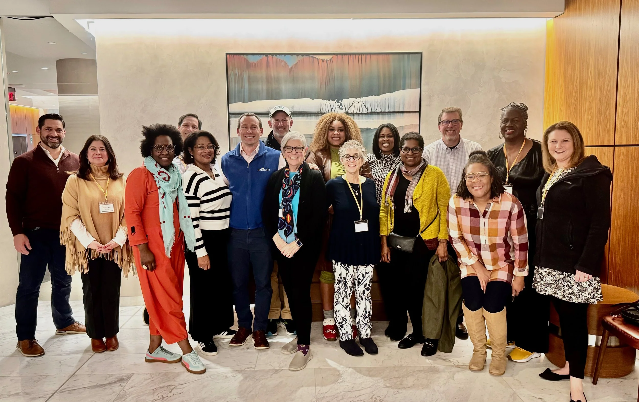 A group of diverse people standing together indoors for a group photo, smiling, with a painting of a waterfall and trees on the wall behind them.