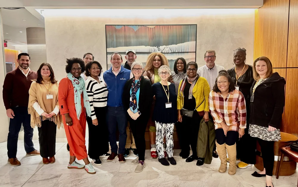 A group of diverse people standing together indoors for a group photo, smiling, with a painting of a waterfall and trees on the wall behind them.