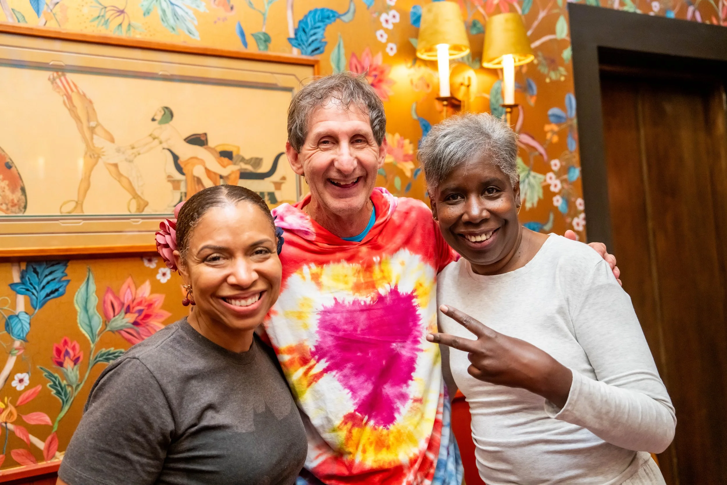 Three smiling people standing close together against a colorful, floral wallpaper background. The person in the middle wears a tie-dye shirt, and the other two women are on either side, smiling warmly.