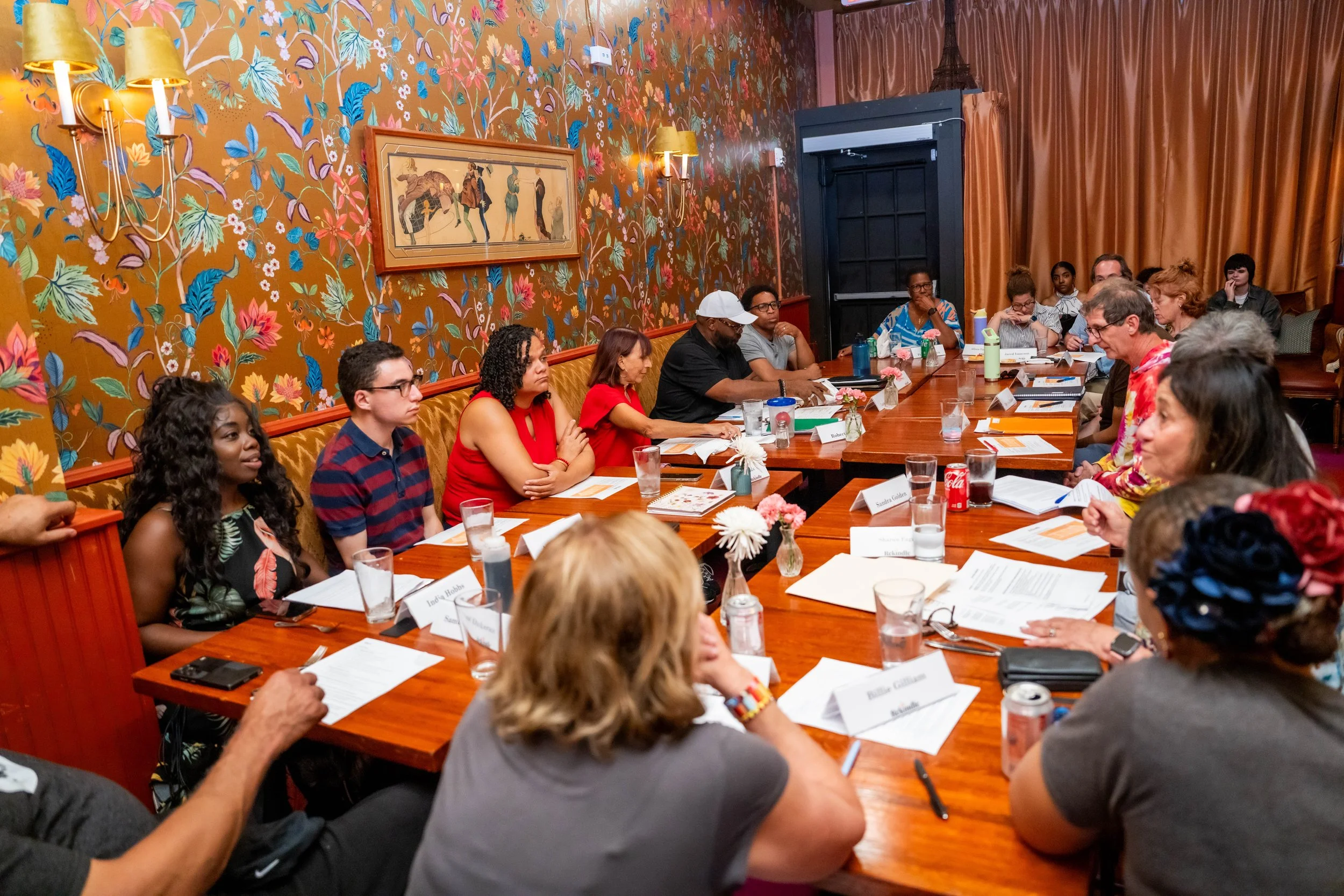 A diverse group of people seated around a large wooden table attending a meeting or conference in a room with floral wallpaper and framed artwork, with some individuals taking notes or listening attentively.