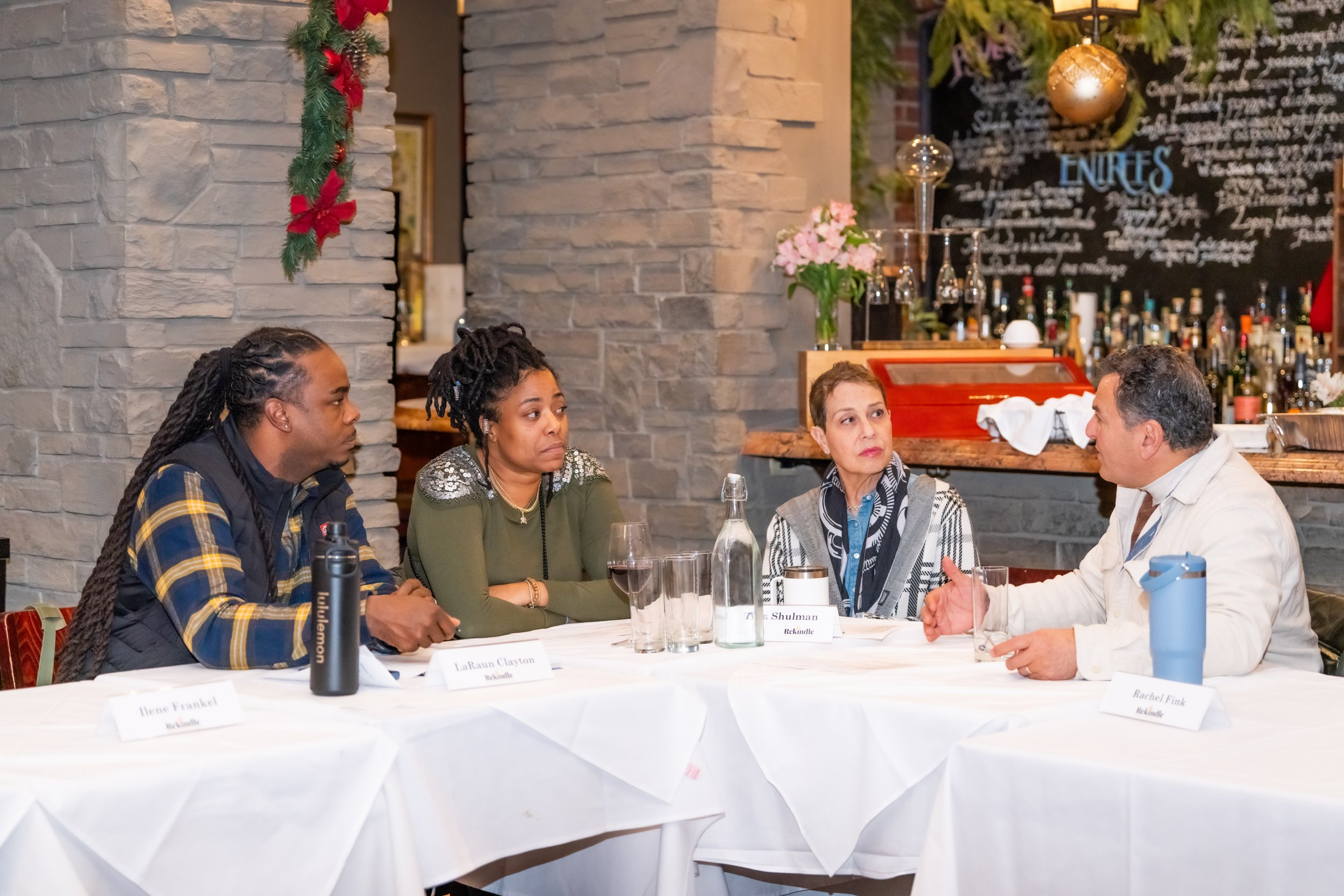A group of four diverse people sitting at a round table engaged in conversation at a formal event. The table is covered with a white tablecloth and has name cards, glasses of water, and a water bottle. The background features a bar area with bottles and glassware, a black chalkboard with handwritten text, and festive holiday decorations including garlands and ornaments.