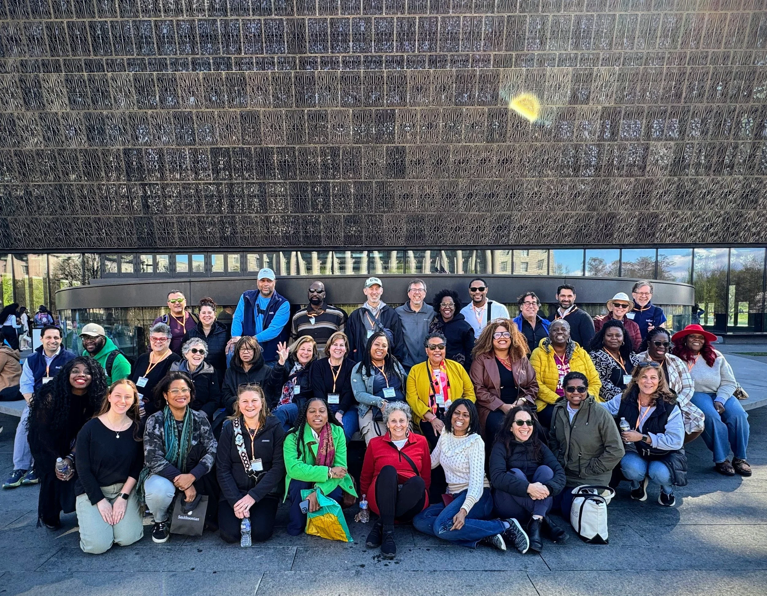 Large group of diverse people gathered outdoors in front of a modern building, smiling and posing for a group photo.